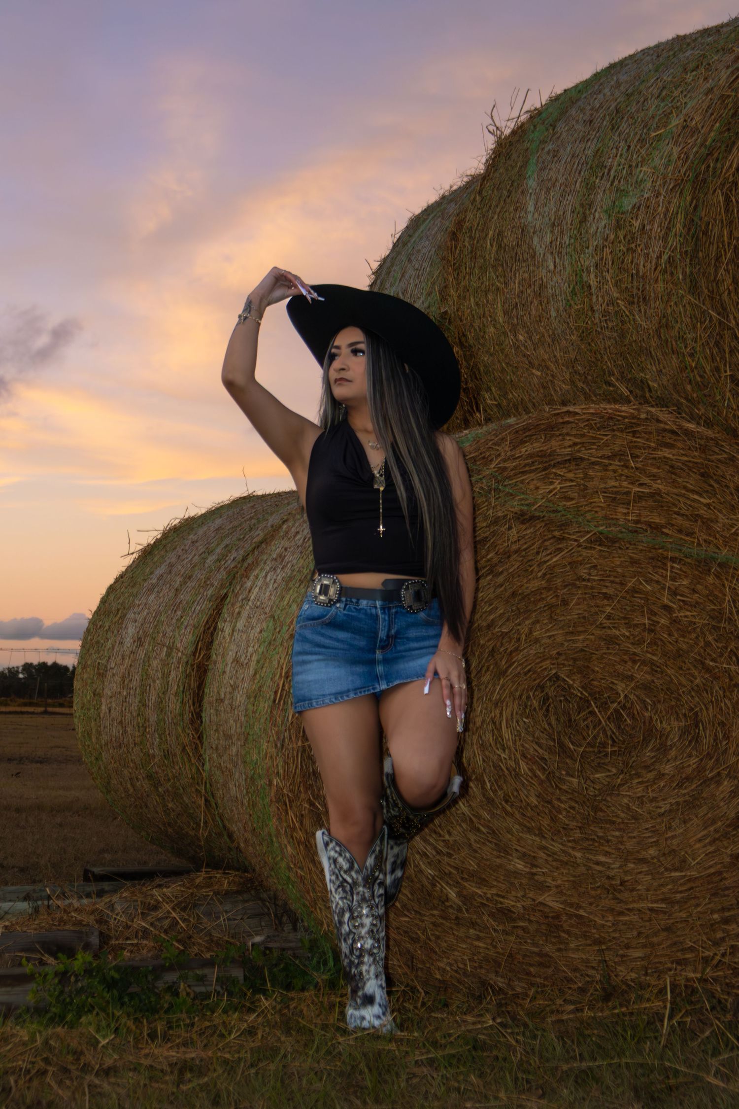 Woman in cowboy hat, denim shorts, and boots leaning against hay bales at sunset on a farm.