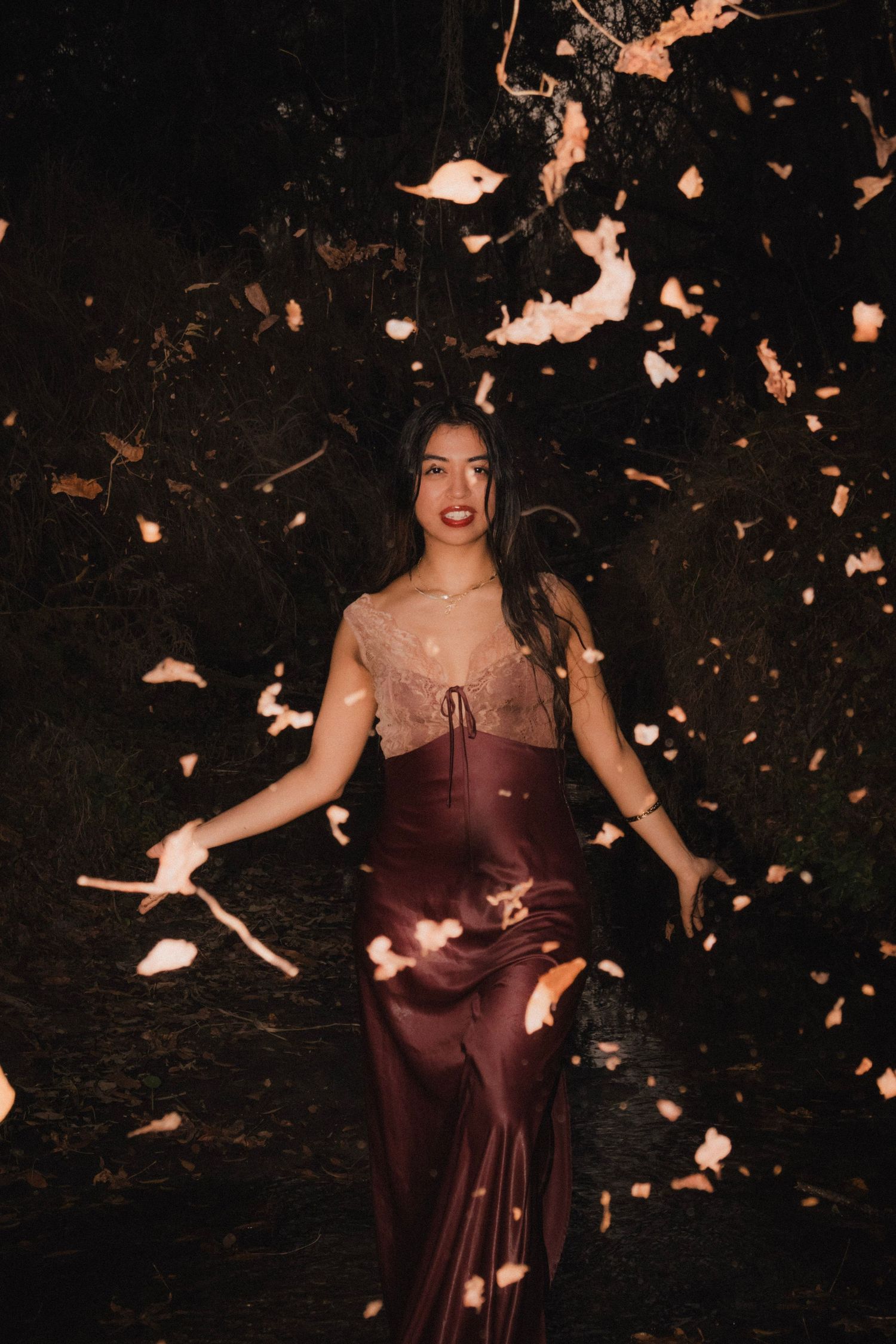 Young woman in burgundy satin gown and lace top posing dramatically with falling petals at night.