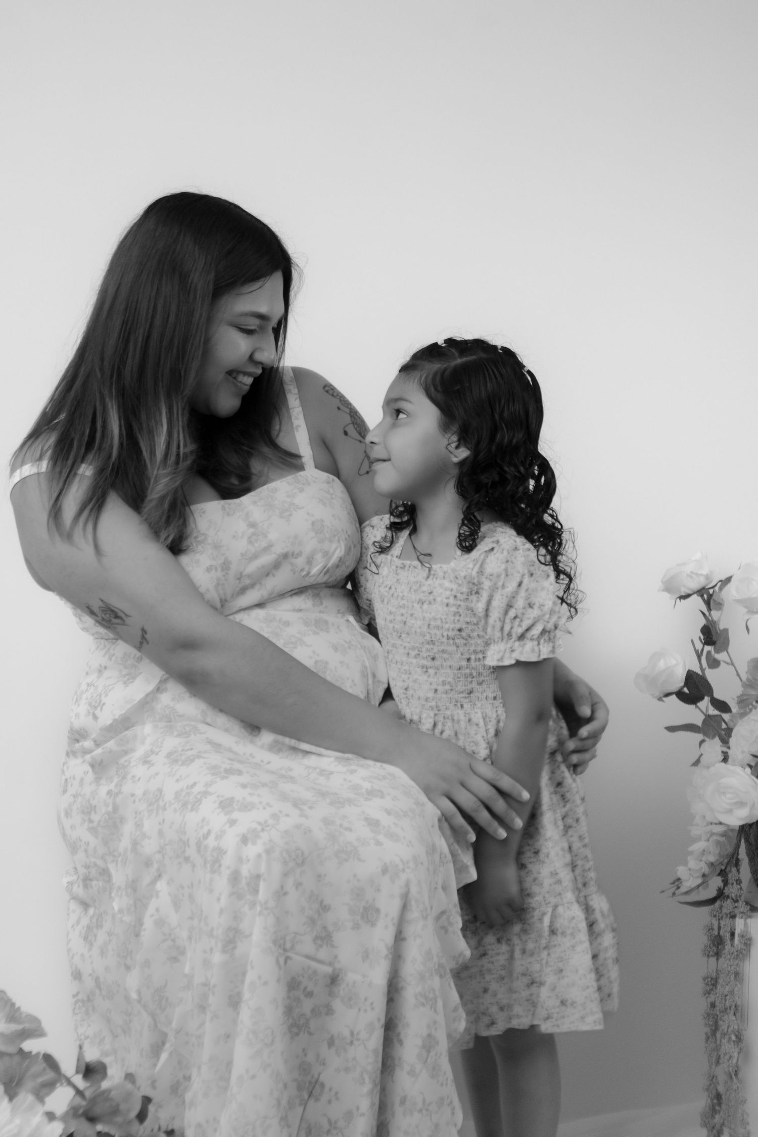 Black and white photo of pregnant woman in floral dress smiling with young curly-haired girl beside flowers.