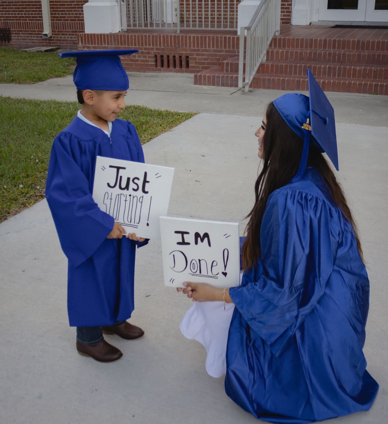 Two graduates in blue caps and gowns holding signs reading Just Starting and Im Done at an outdoor ceremony.