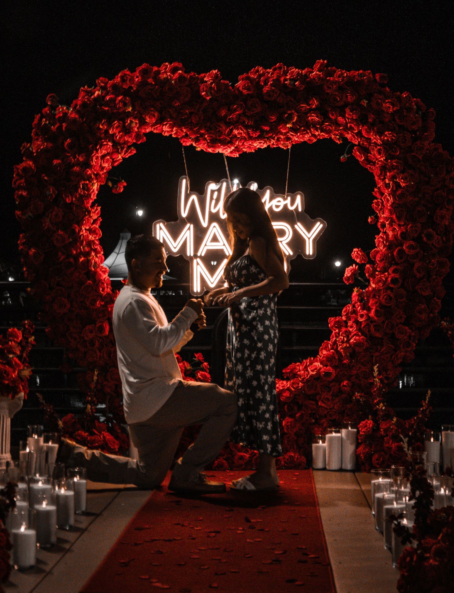 A romantic marriage proposal scene with red rose heart arch and glowing neon Will You Marry Me sign.