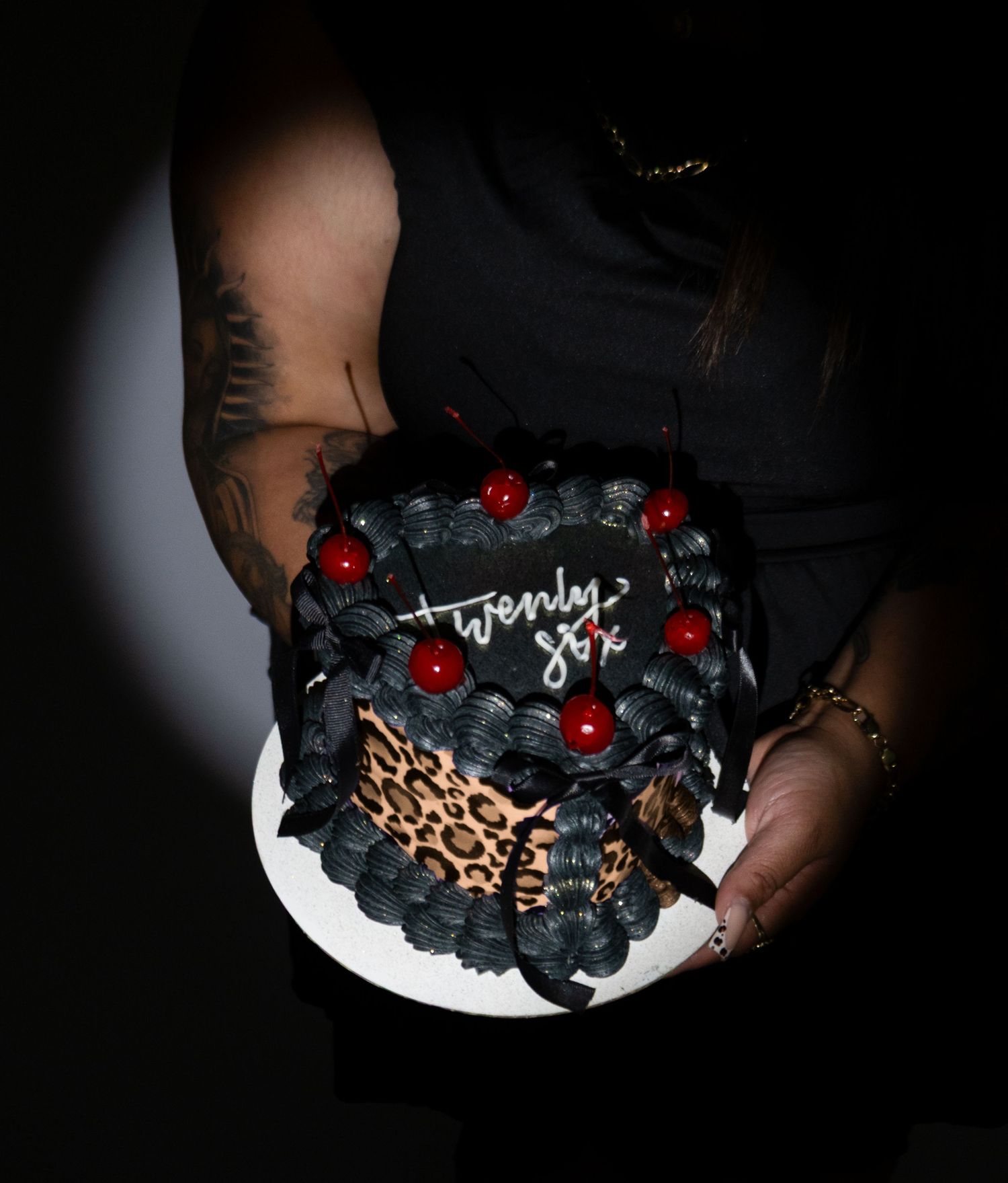 Tattooed person holding a dark black birthday cake decorated with red cherries and leopard print reading twenty six.