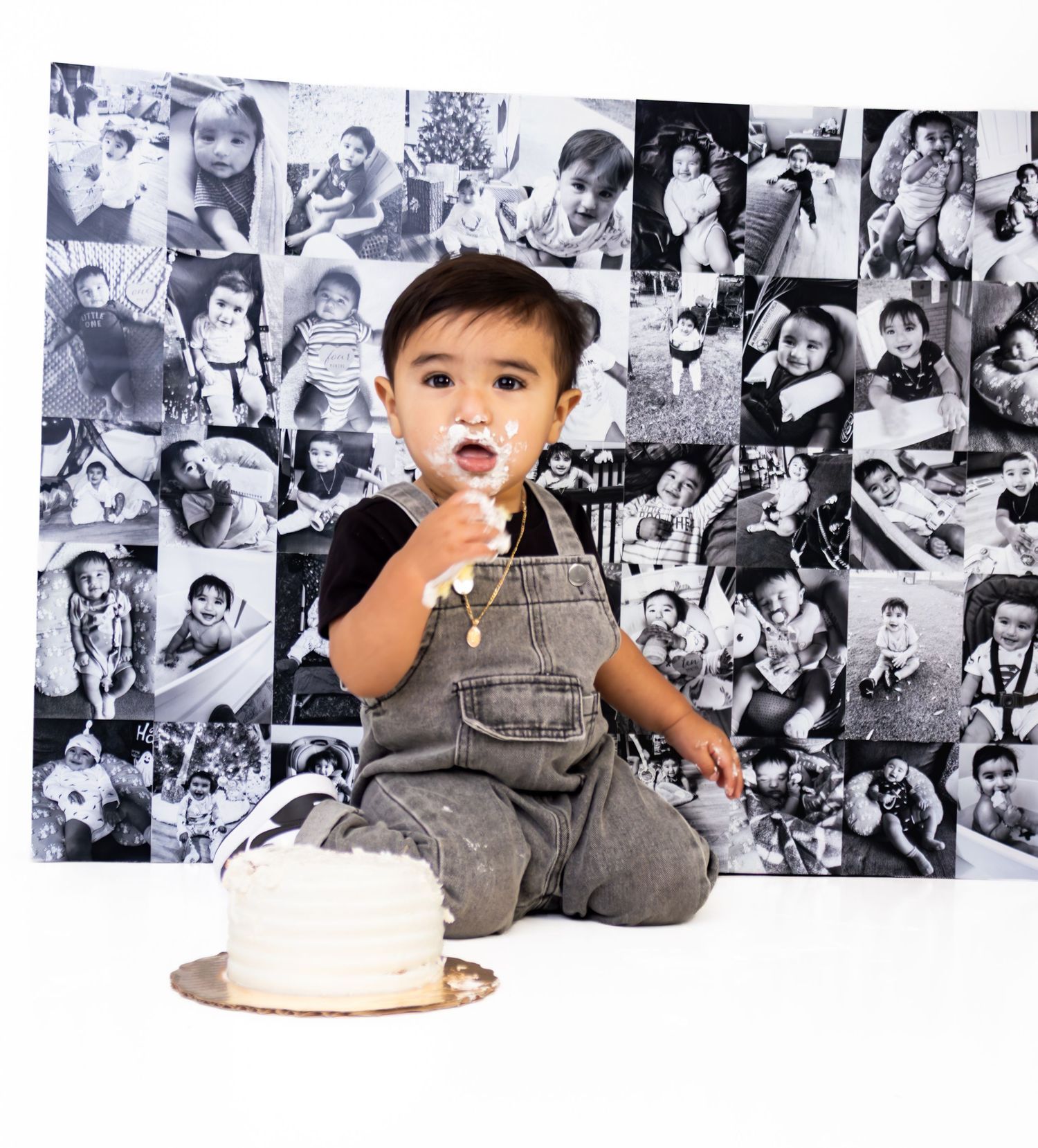 Toddler boy in denim overalls enjoys first birthday cake smash in front of photo collage backdrop.