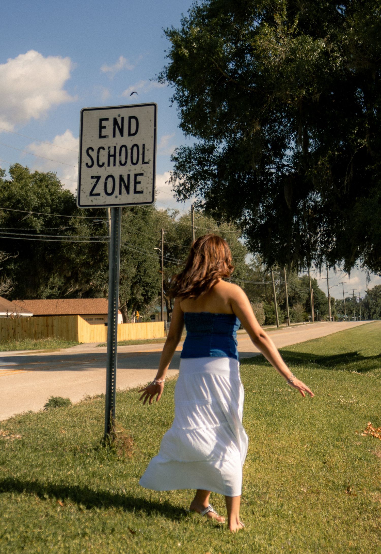 A woman in a blue top and white skirt stands near an End School Zone sign on a sunny day.