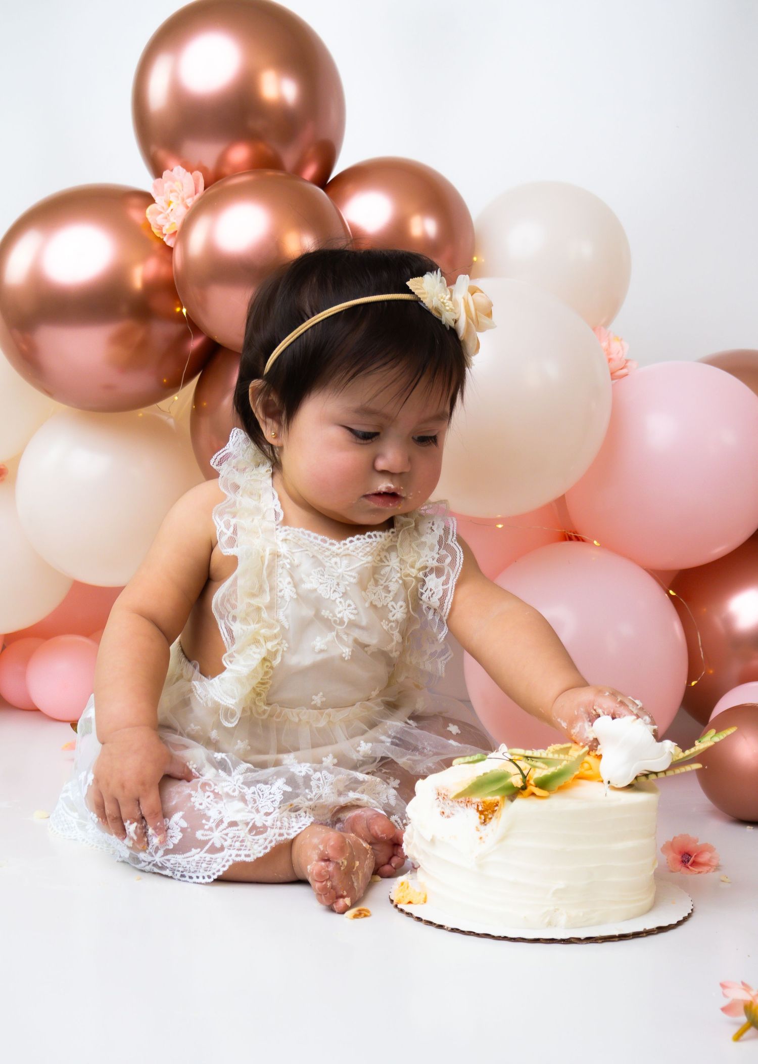 One year old baby girl in white lace dress smashing birthday cake surrounded by pink and rose gold balloons.
