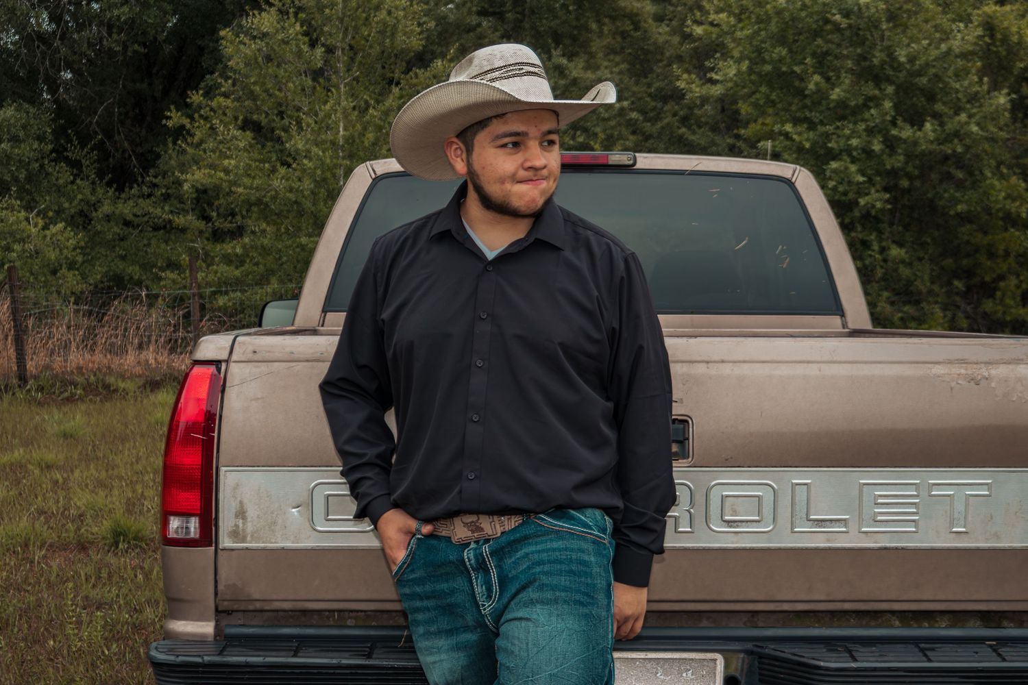 Young man in cowboy hat and dark shirt leaning against a Chevrolet truck tailgate in a rural setting.