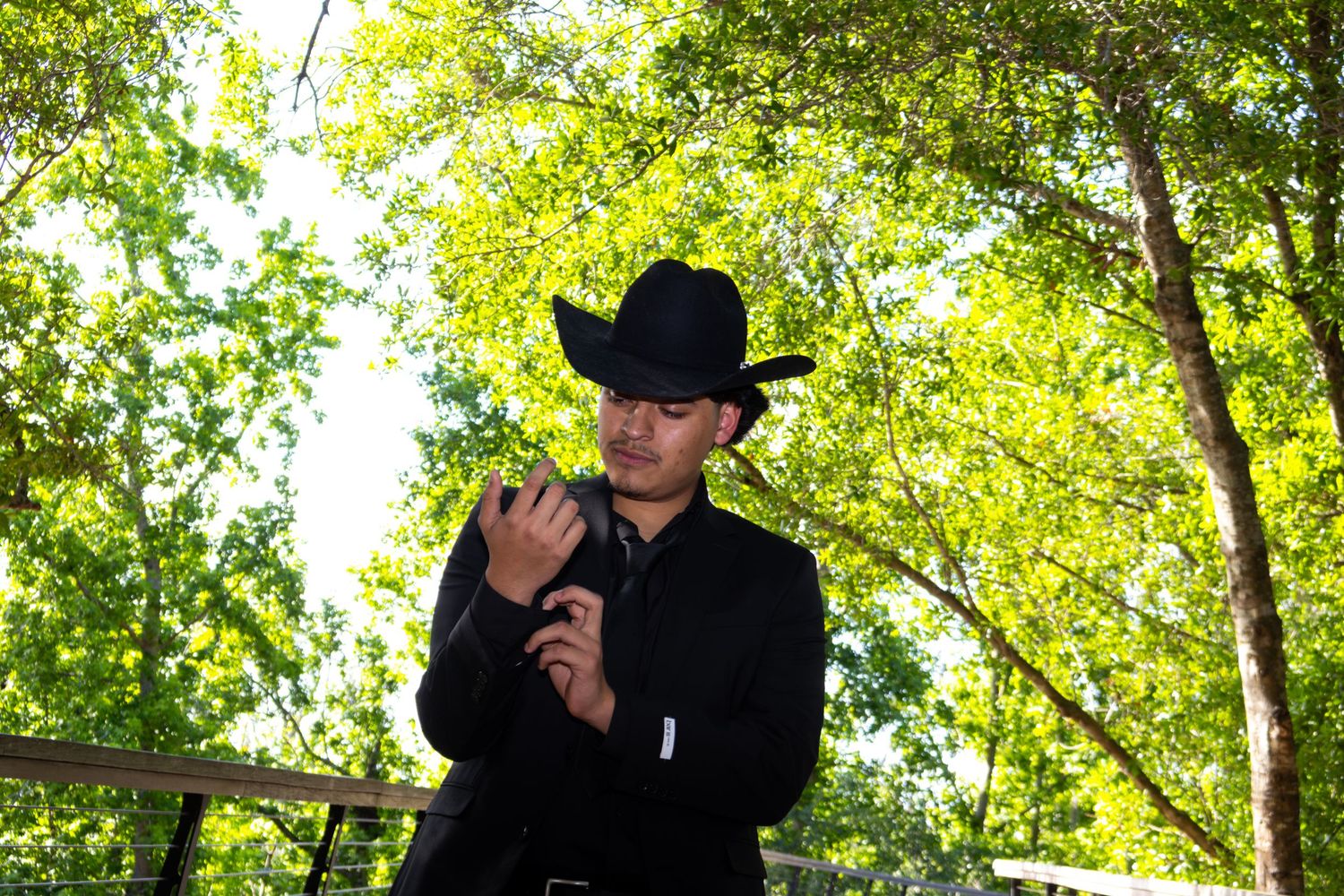 A young man in black shirt and cowboy hat adjusts his sleeve outdoors among green trees.