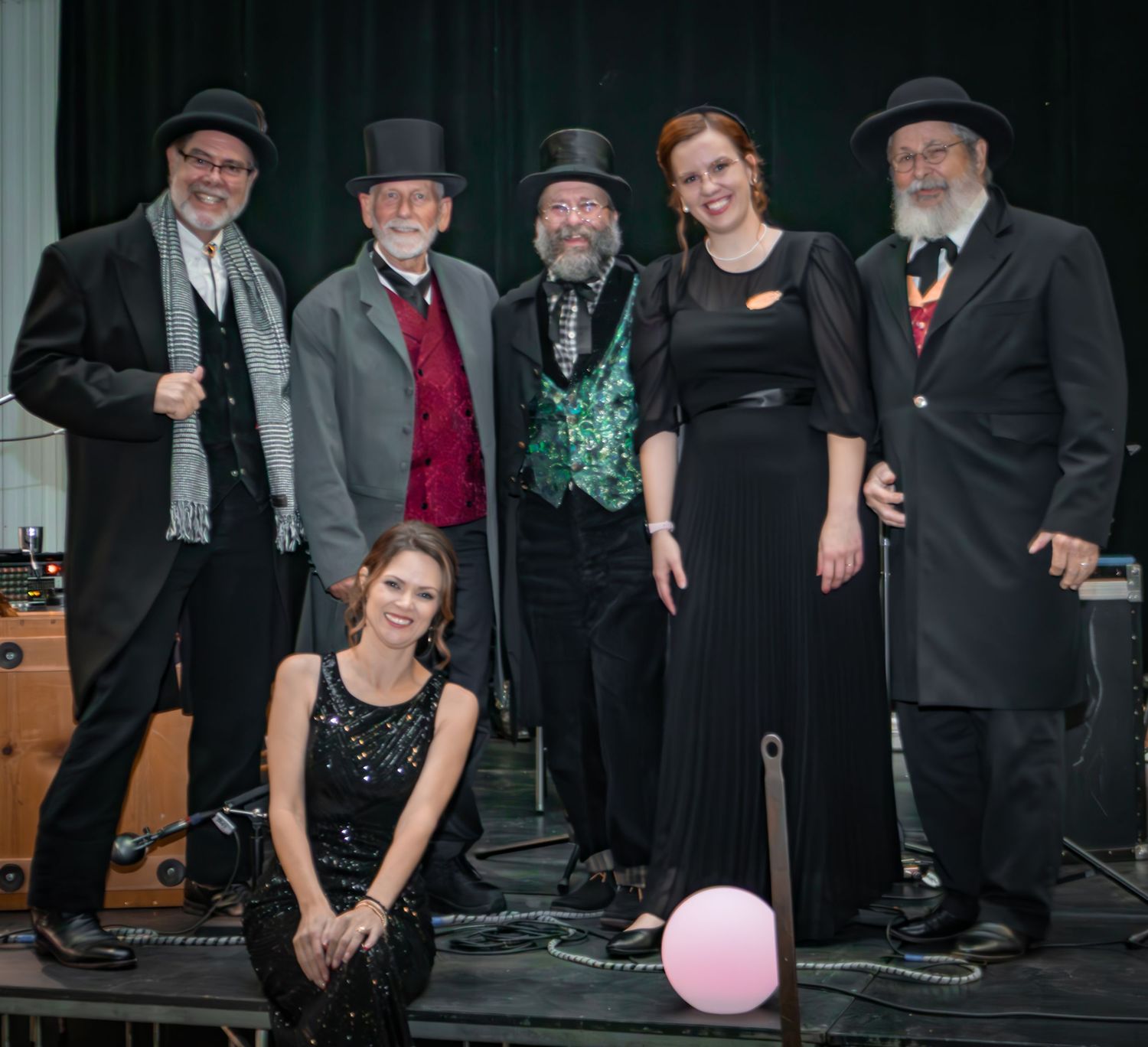 A six-member Victorian-themed performance band poses together on stage in period costumes at a formal event.