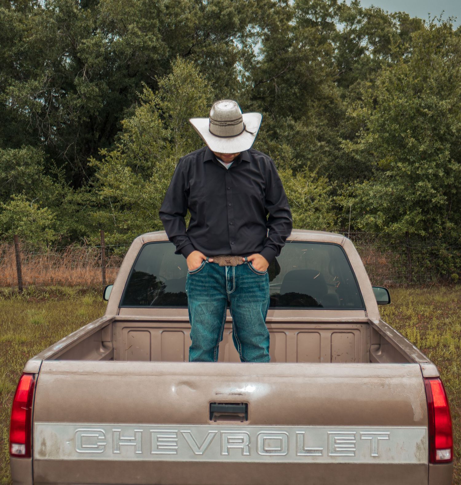 Man in cowboy hat and jeans leaning against a tan Chevrolet pickup truck bed outdoors.