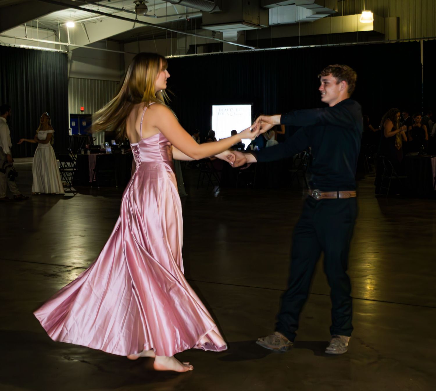 A couple dances elegantly, the woman wearing a flowing pink satin gown at a formal ballroom event.