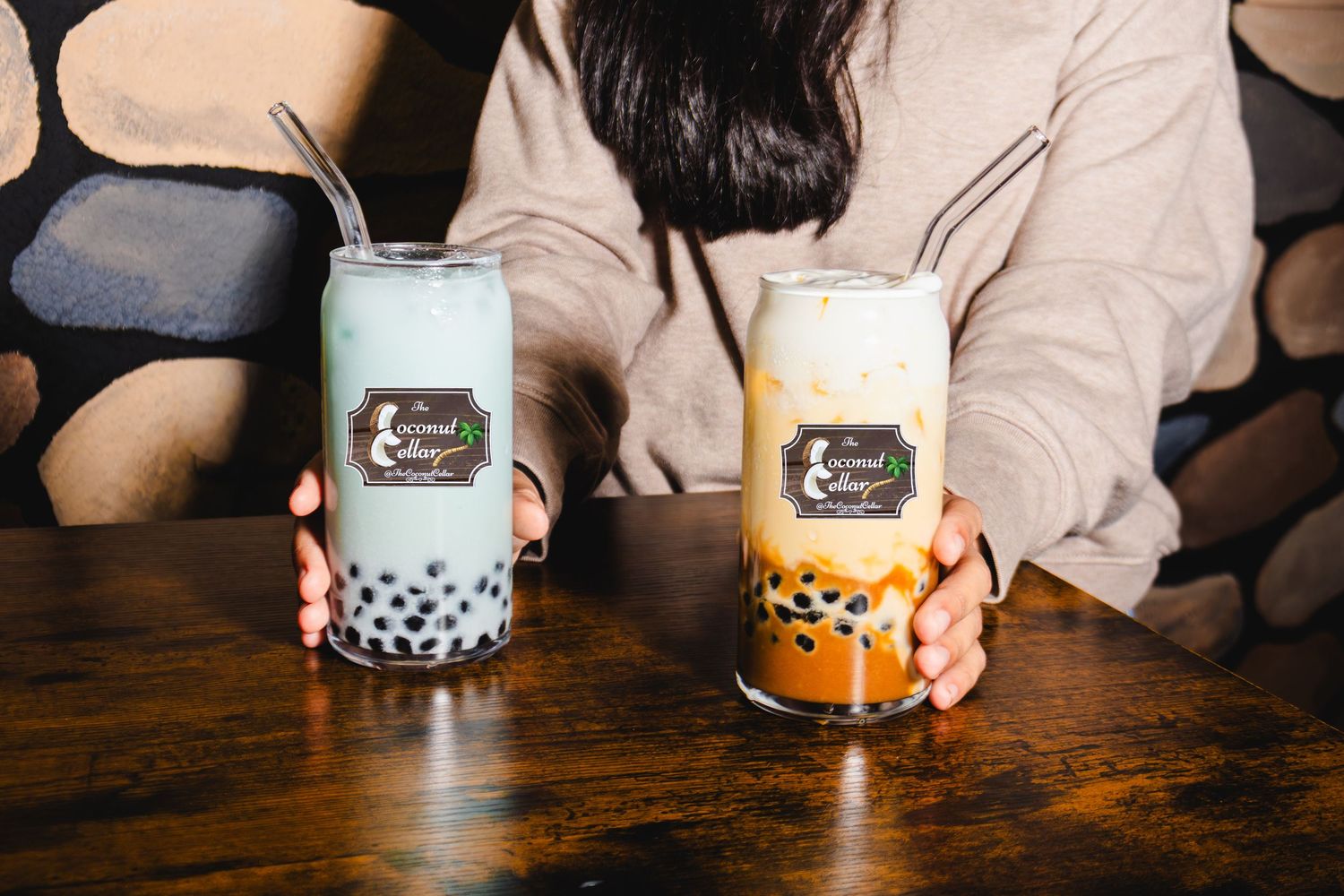 Person holding two Coconut Cellar boba drinks with glass straws and black tapioca pearls on wooden table.