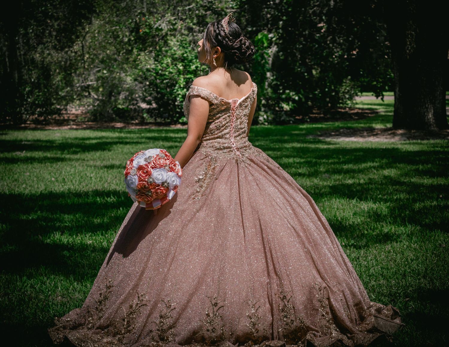 A young woman in a rose gold embroidered quinceañera gown holds a bouquet while posing in a lush garden.