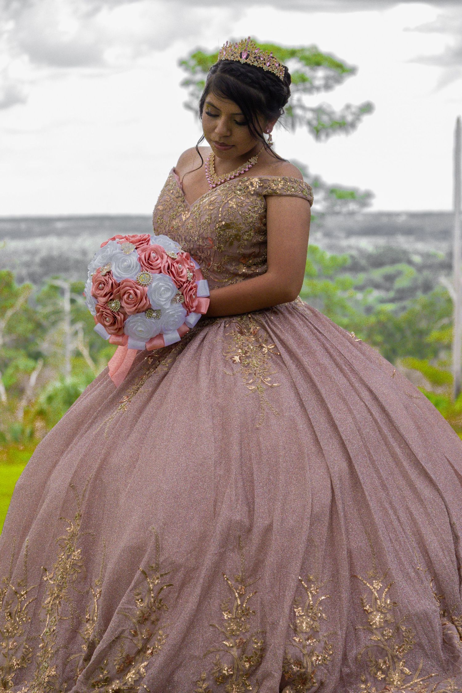 A young woman in a rose gold quinceañera ball gown with gold embroidery holds a pink bouquet outdoors.