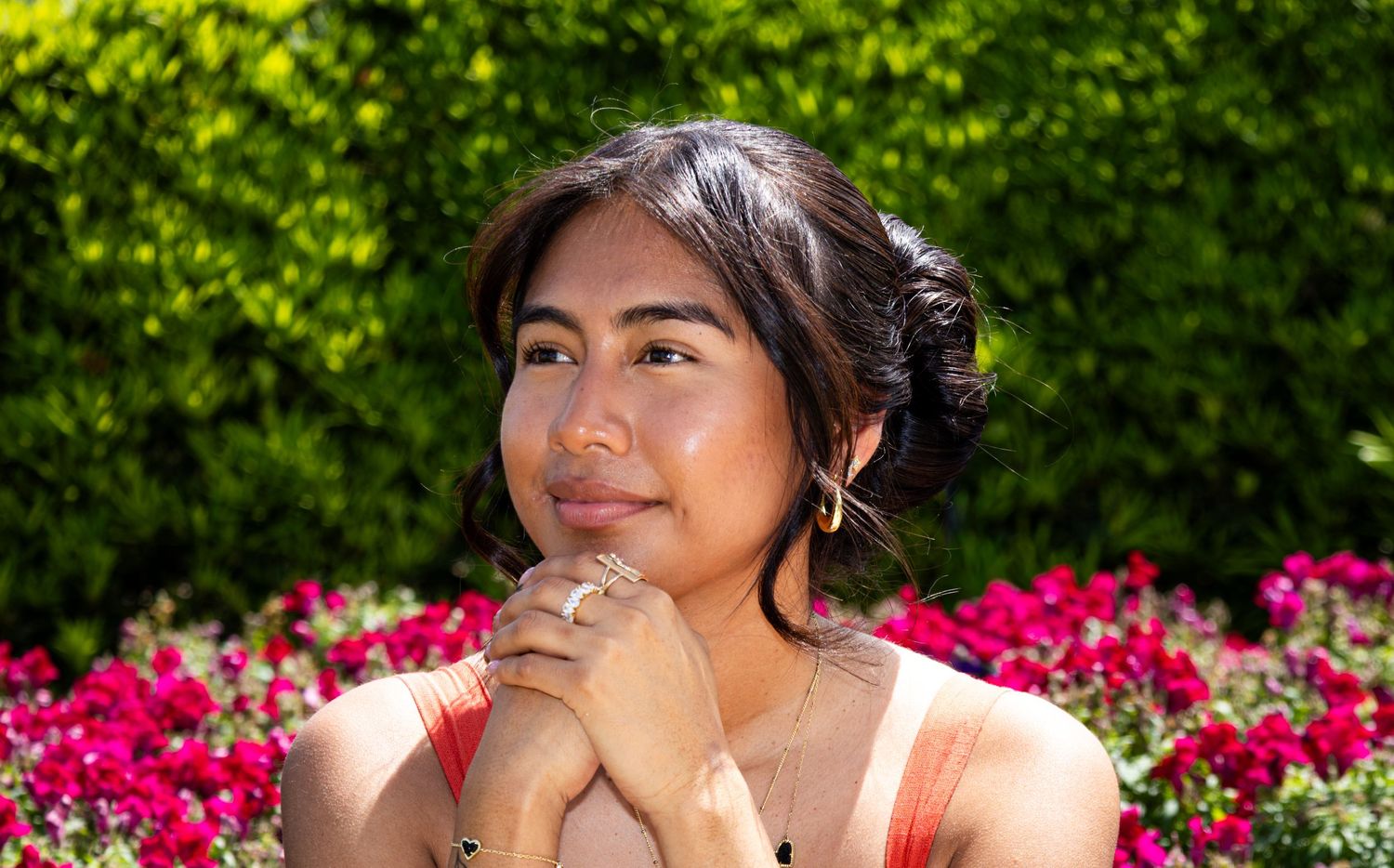 Young woman with updo smiling softly in front of vibrant pink flower garden.