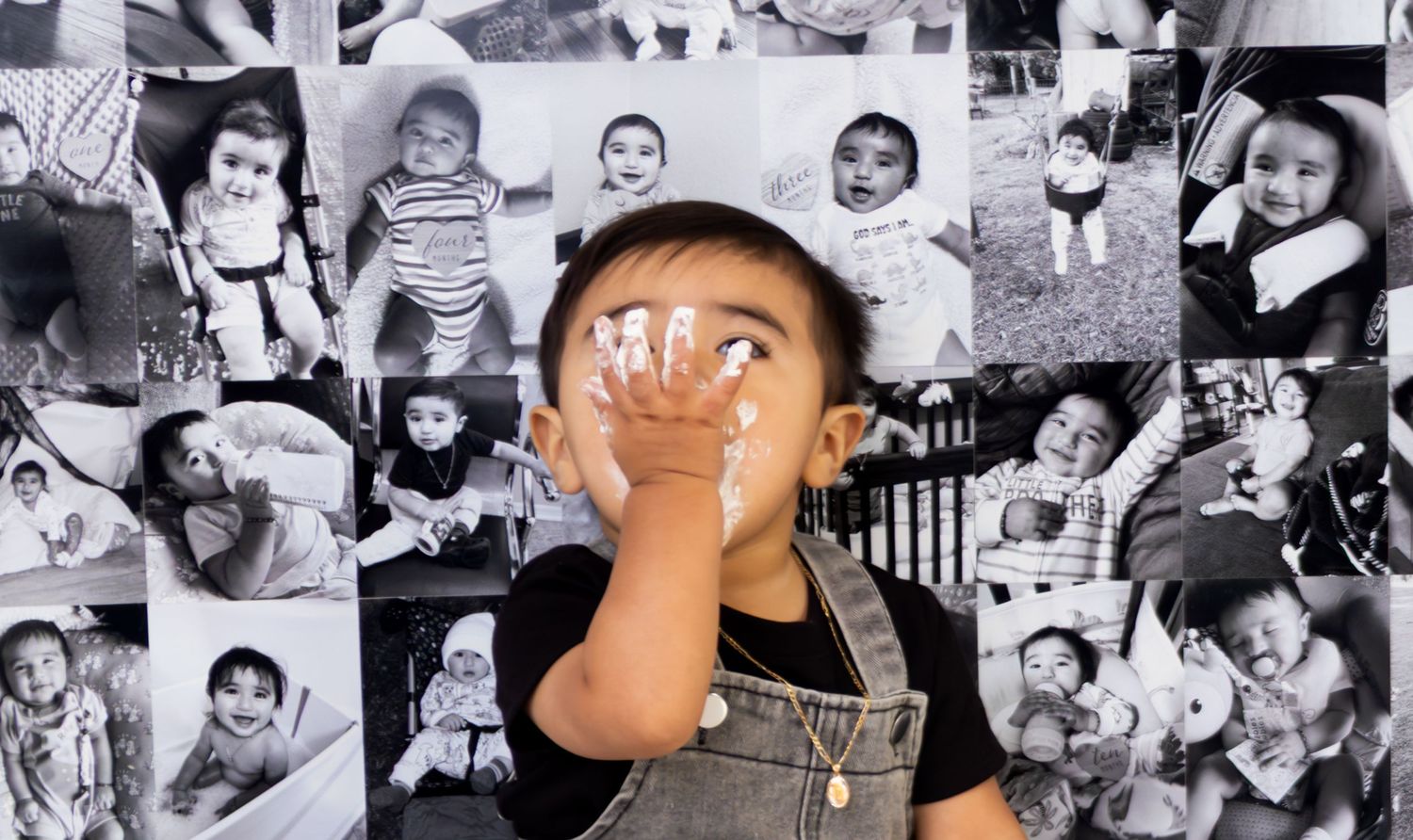 A baby covers their face with their hand in front of a black and white photo collage backdrop.