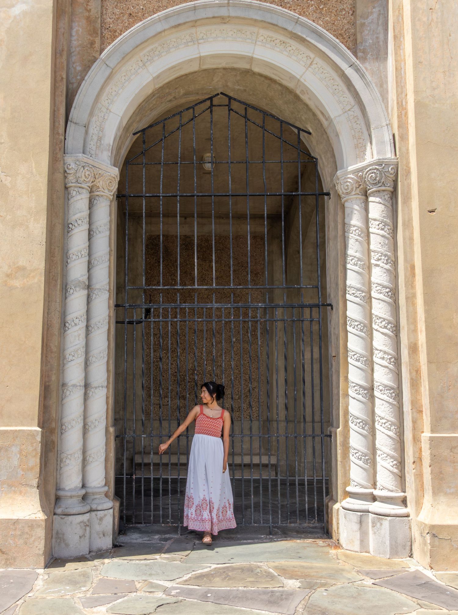 Young woman in red top and white skirt standing in ornate stone archway with iron gate.