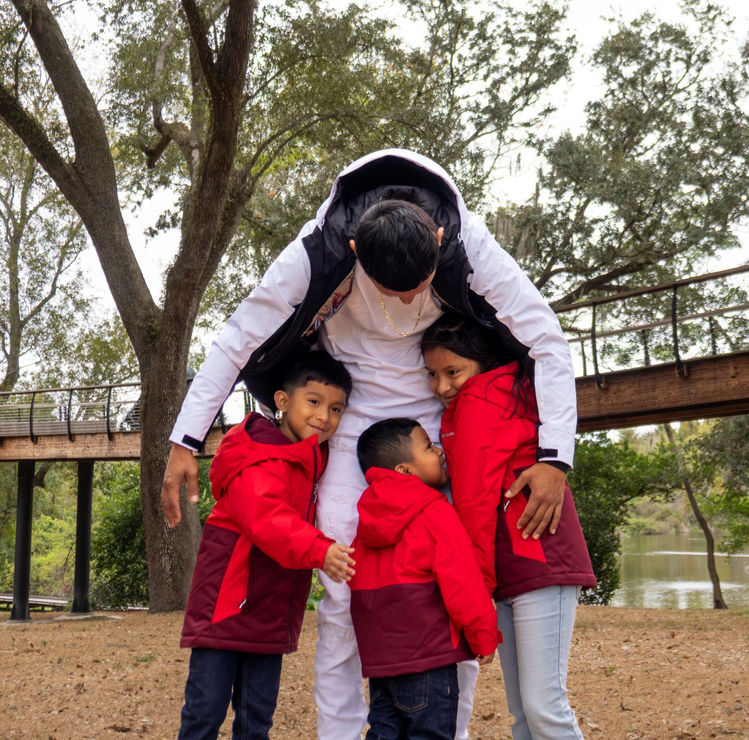 Adult in white hoodie hugging three children in red jackets outdoors near trees and a wooden bridge.
