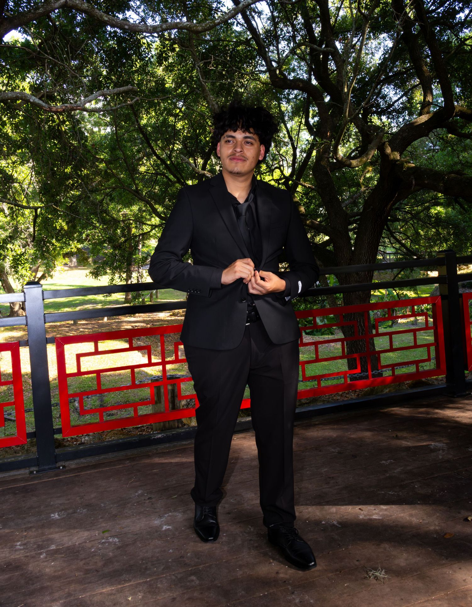 Young man in black suit posing outdoors near red decorative fence with lush green trees in background.