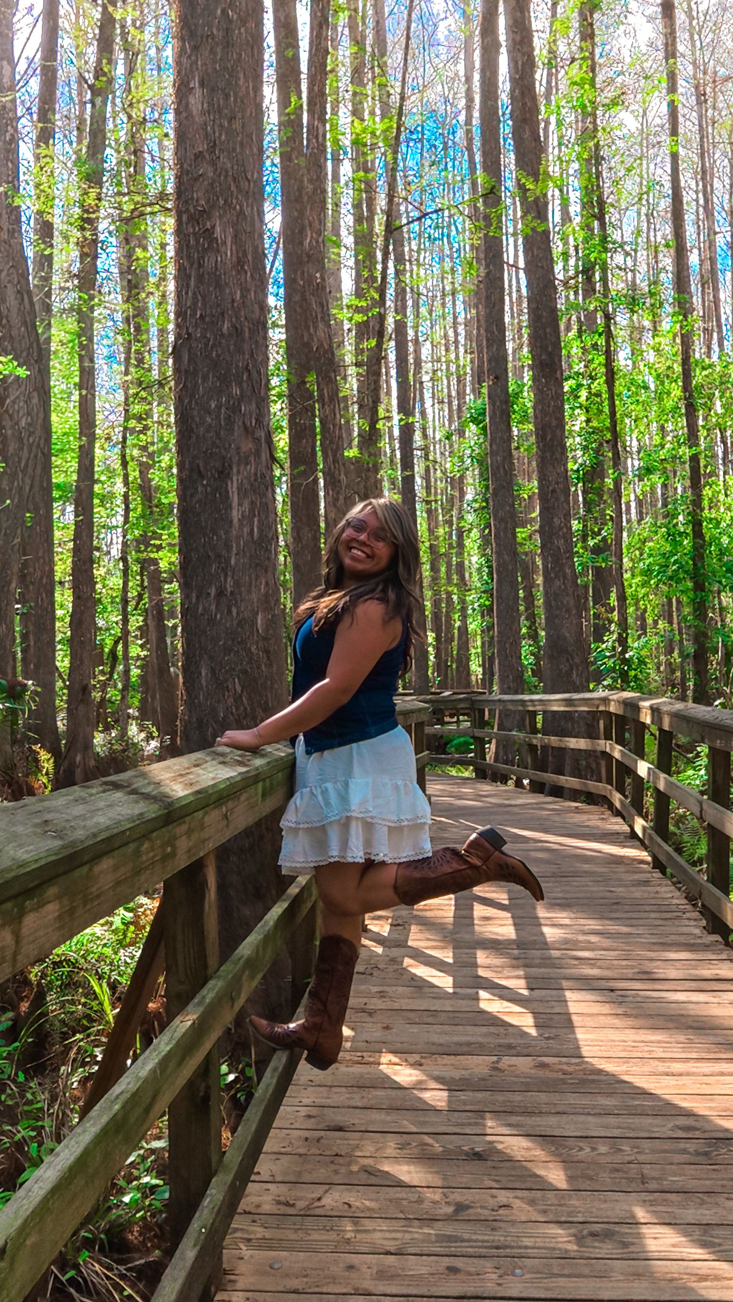 Cheerful woman in white skirt and boots poses on wooden forest boardwalk surrounded by tall trees.