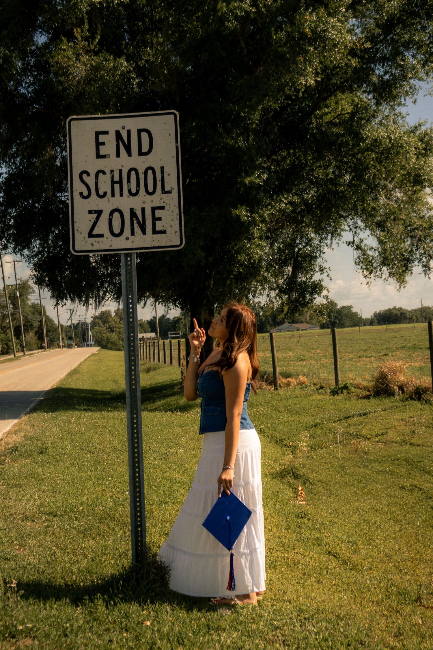 A graduate poses beside an End School Zone sign holding her blue cap, symbolizing the completion of her education.