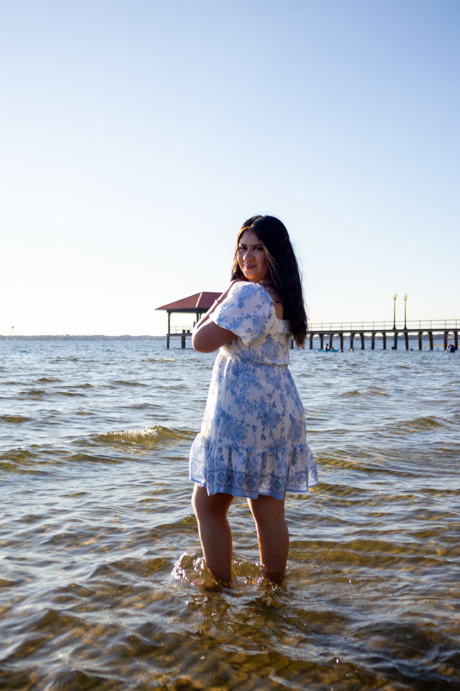Young woman in floral sundress standing in shallow ocean water near a pier on a sunny day.