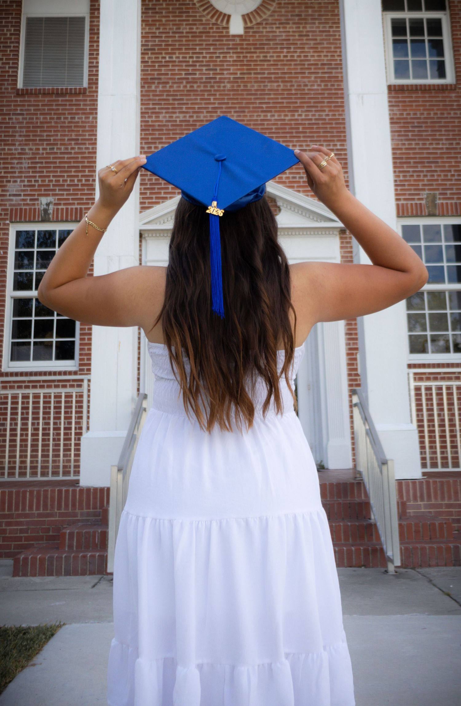 A young woman in a white dress adjusts her blue graduation cap while standing in front of a brick university building.