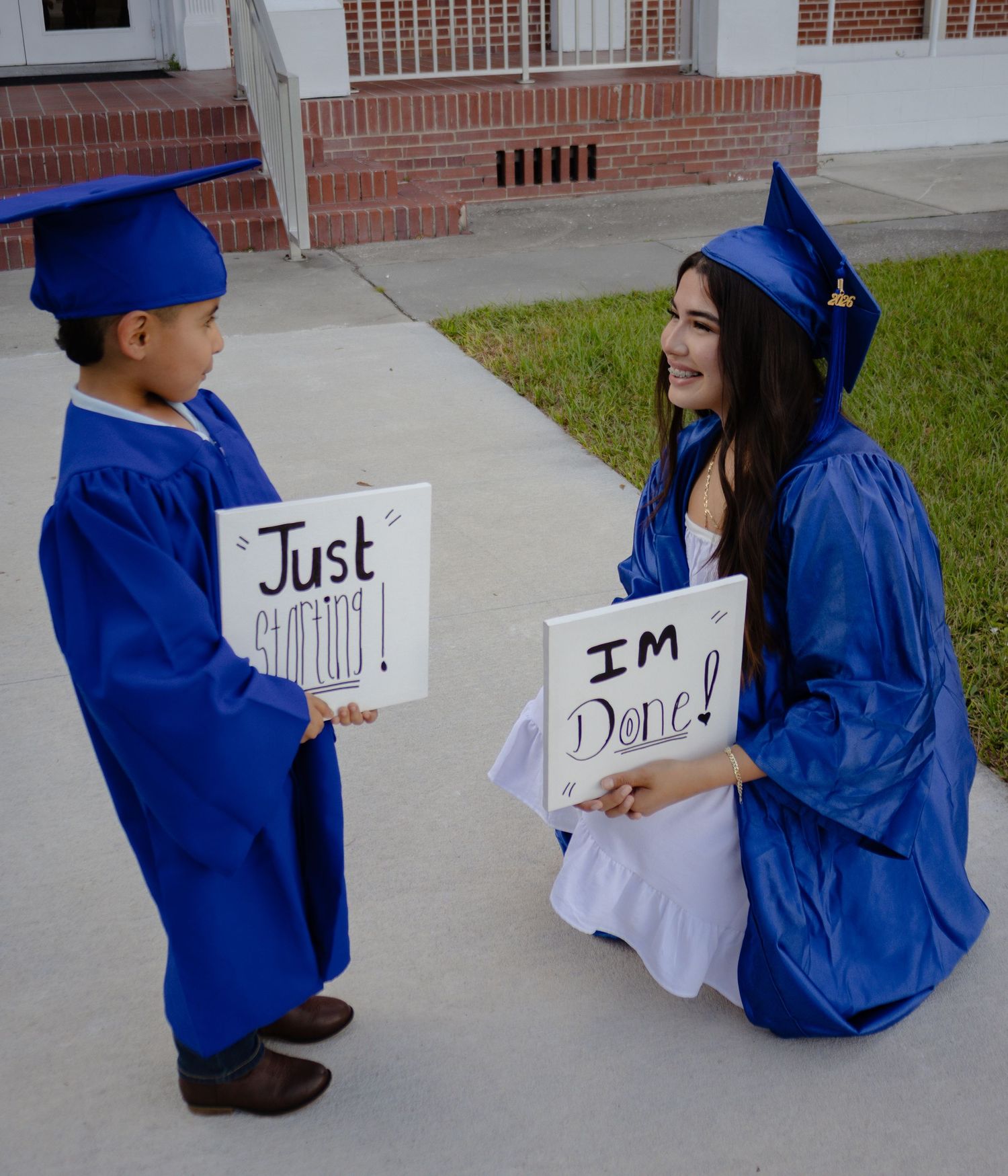 A young boy and teenage girl in blue graduation caps and gowns hold signs reading Just Starting and Im Done.