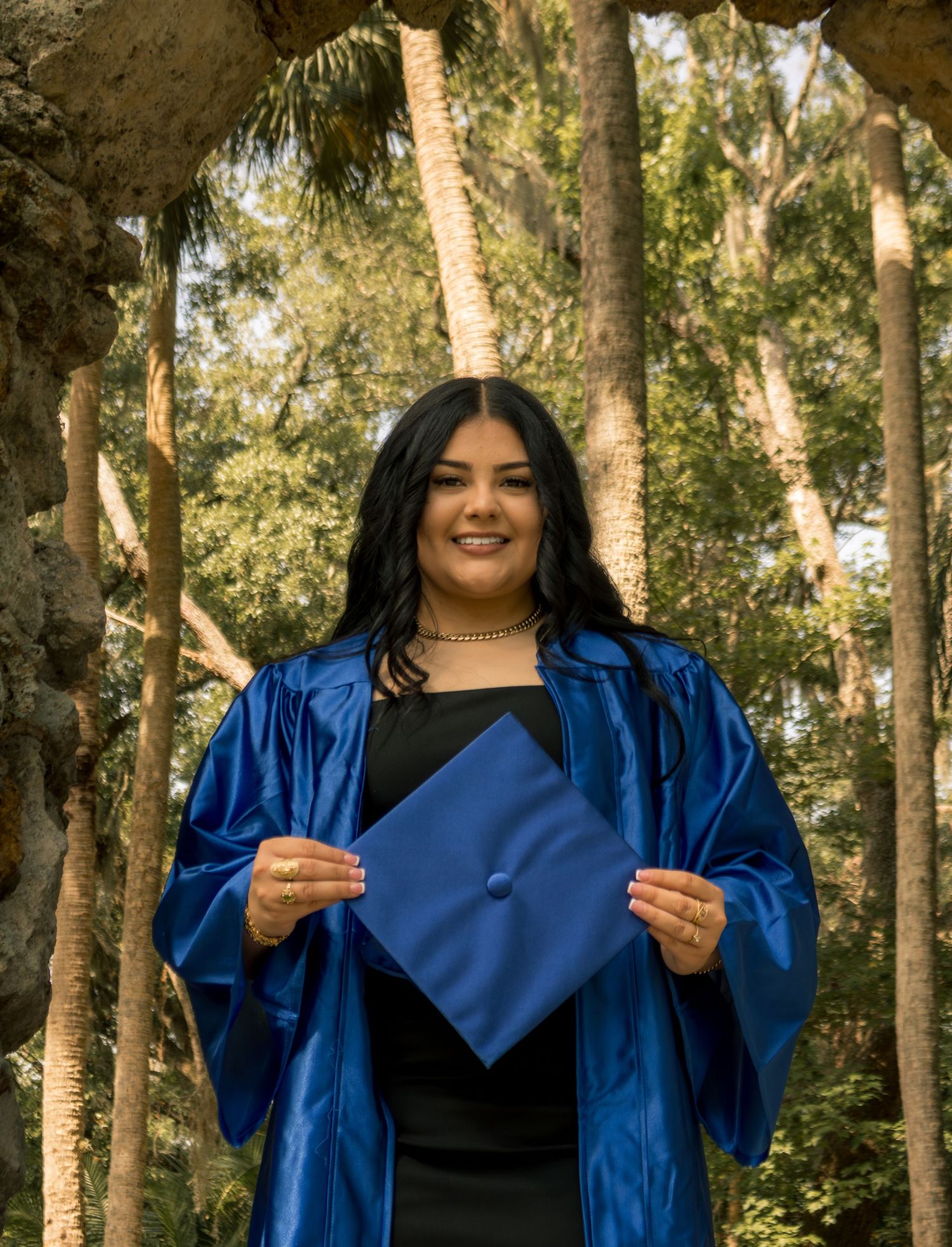 A smiling graduate in a blue cap and gown holds her mortarboard in front of lush green trees.