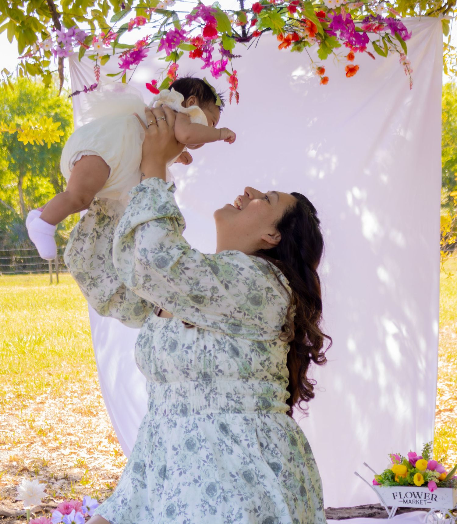 Mother in floral dress lifting laughing baby girl under colorful flower arch in outdoor photo session.