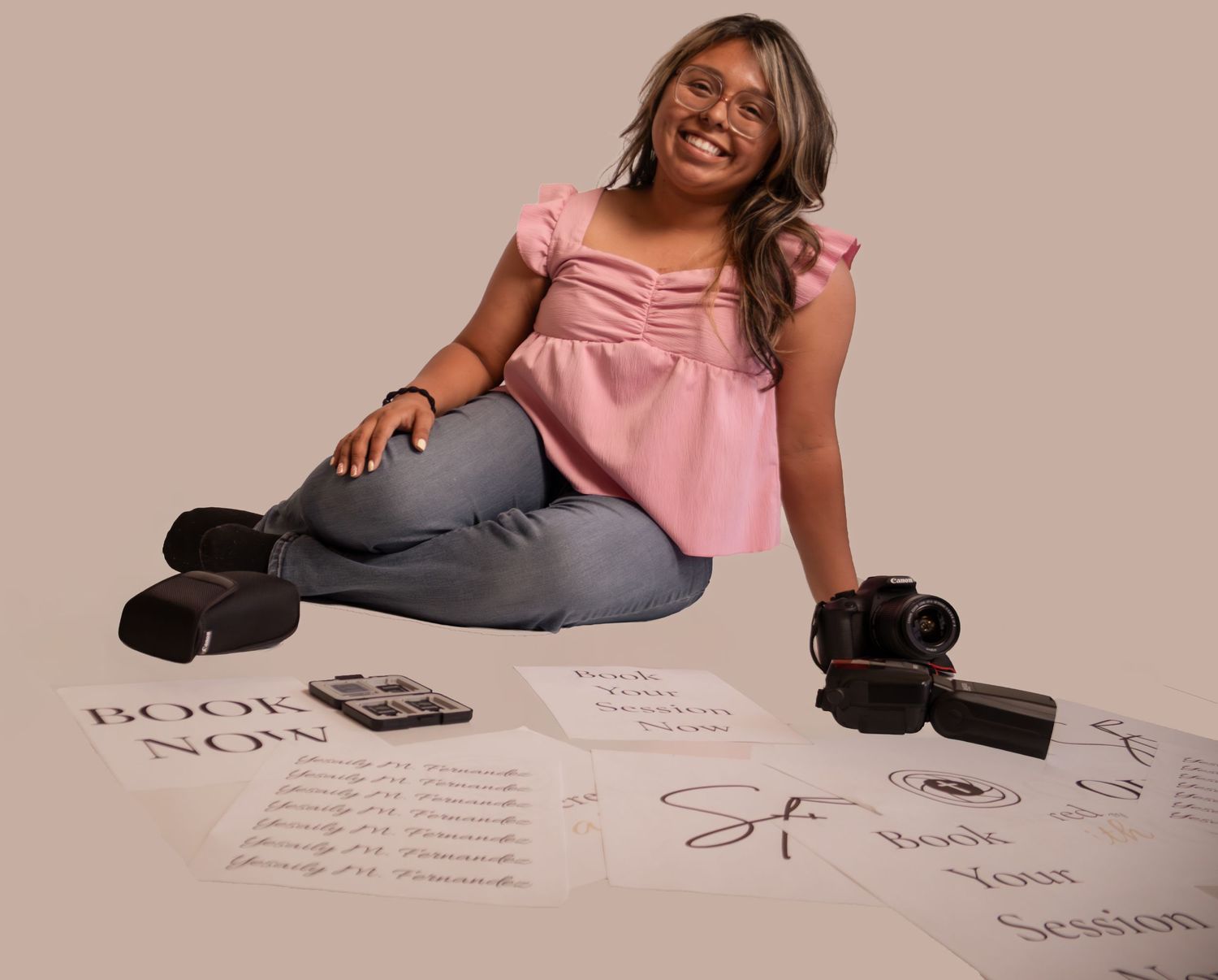 Smiling woman in pink top sits on floor surrounded by booking forms, cameras, and business papers on beige background.