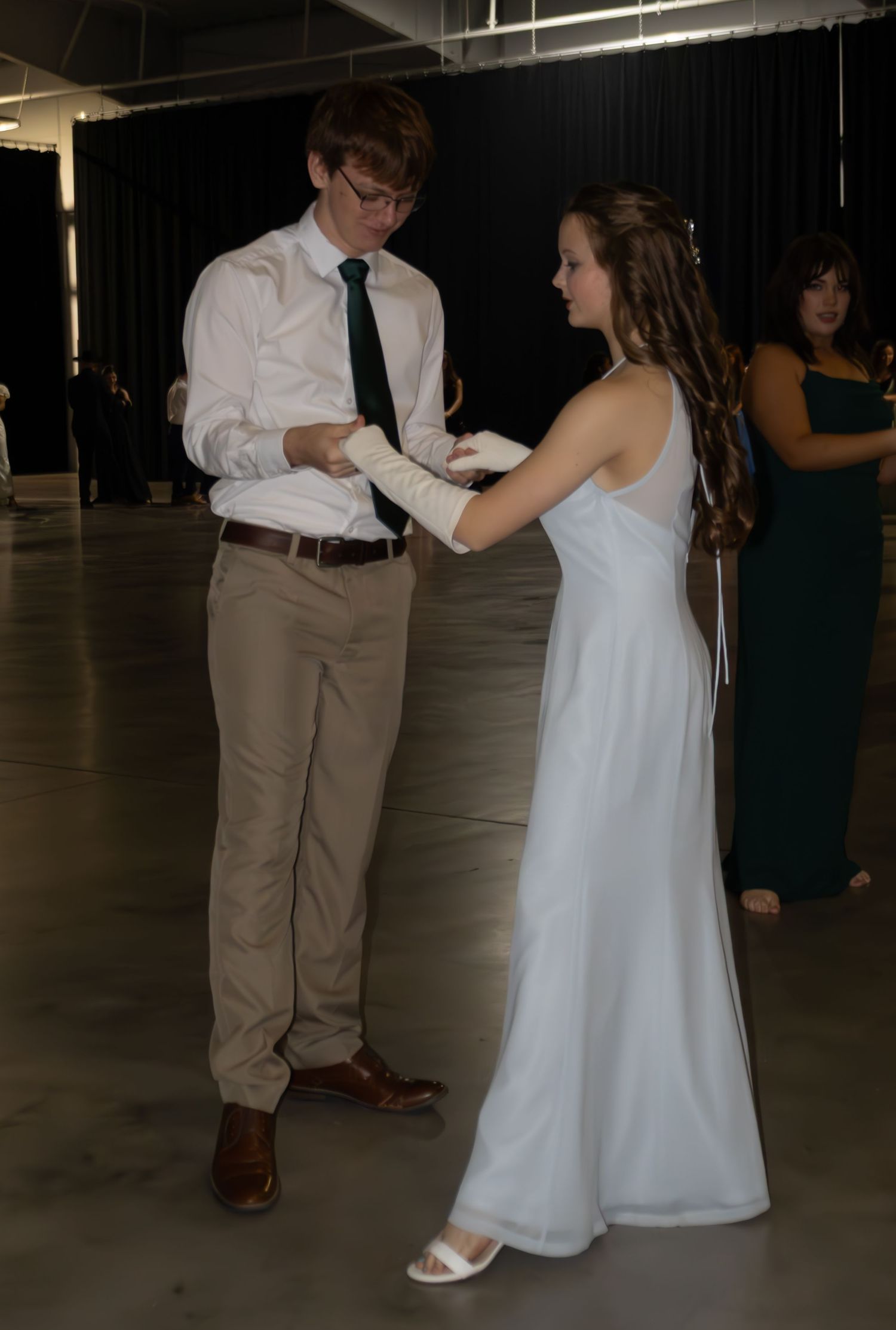 A young man in formal attire places white gloves on a girl in a white gown at a formal dance event.
