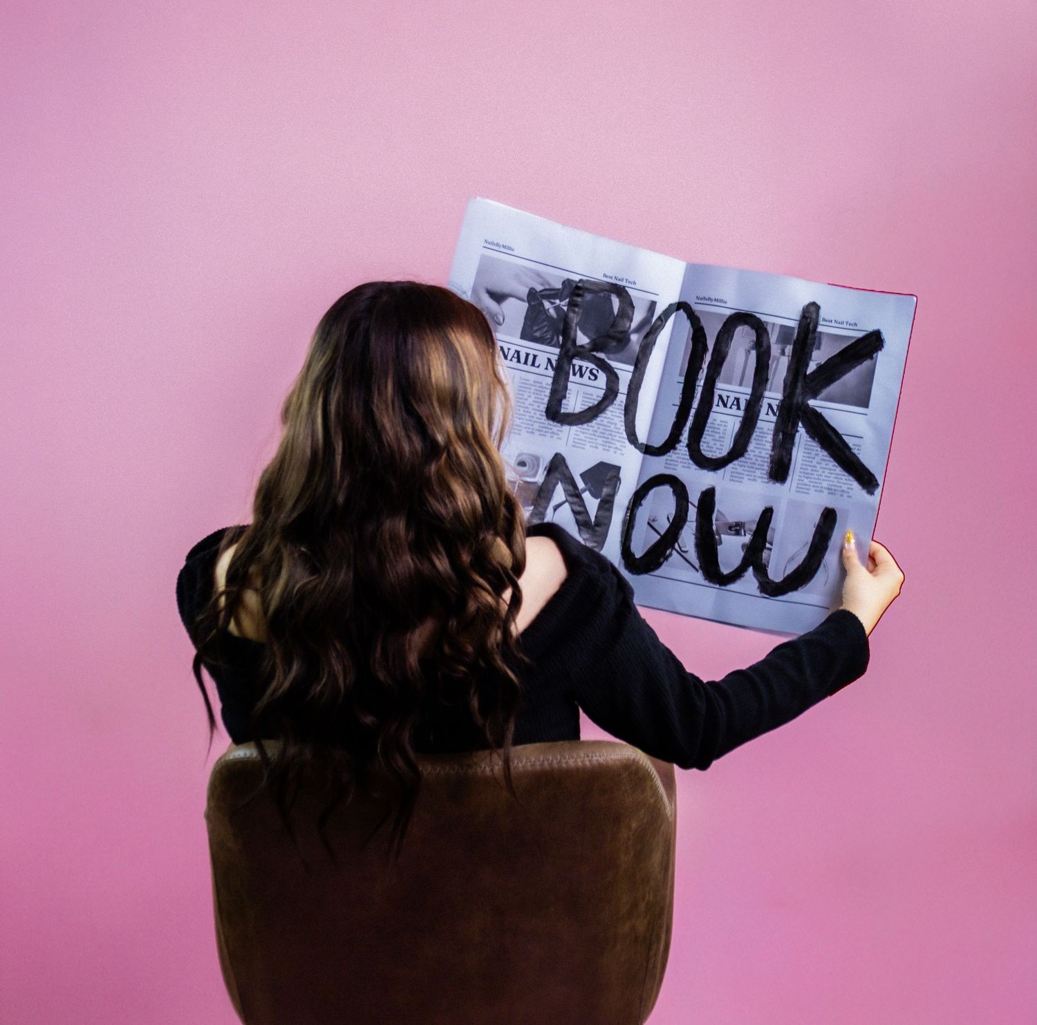 Woman with long brown hair sitting in chair holding open newspaper with Book Now written on pink background.