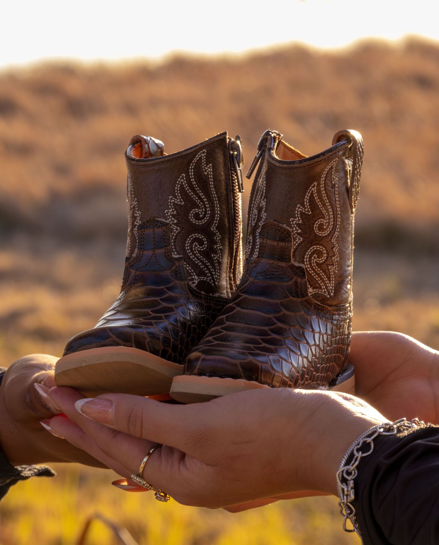 Hands holding tiny brown snakeskin cowboy boots in golden desert sunlight.