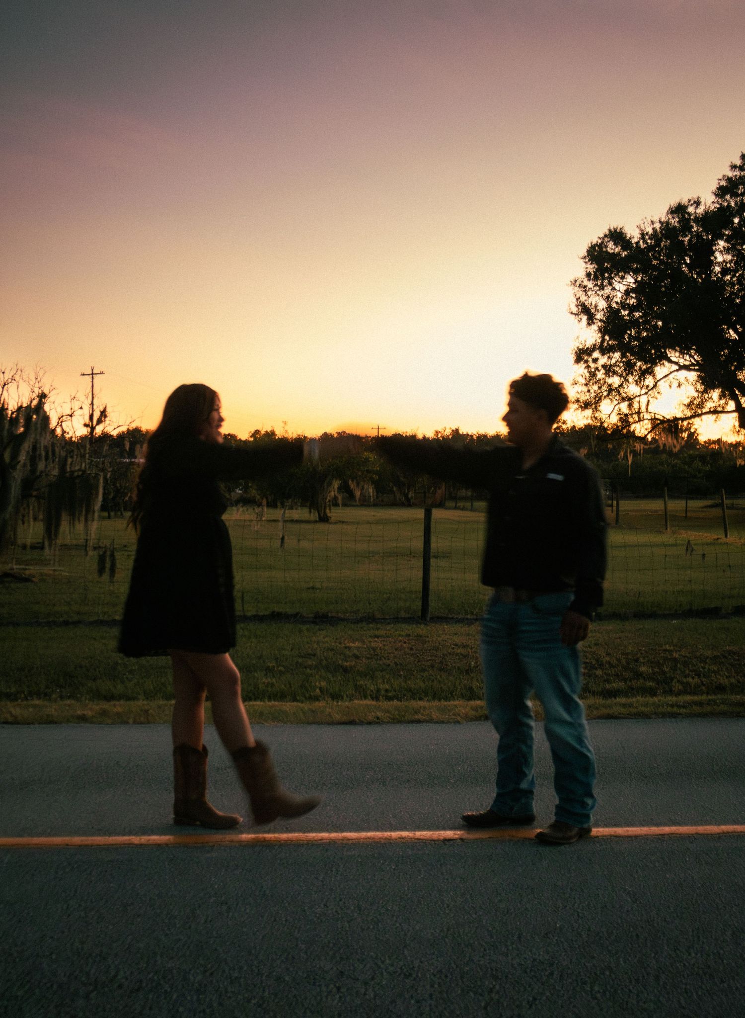 Couple silhouetted against a golden sunset standing on a rural road with fields and trees behind them.