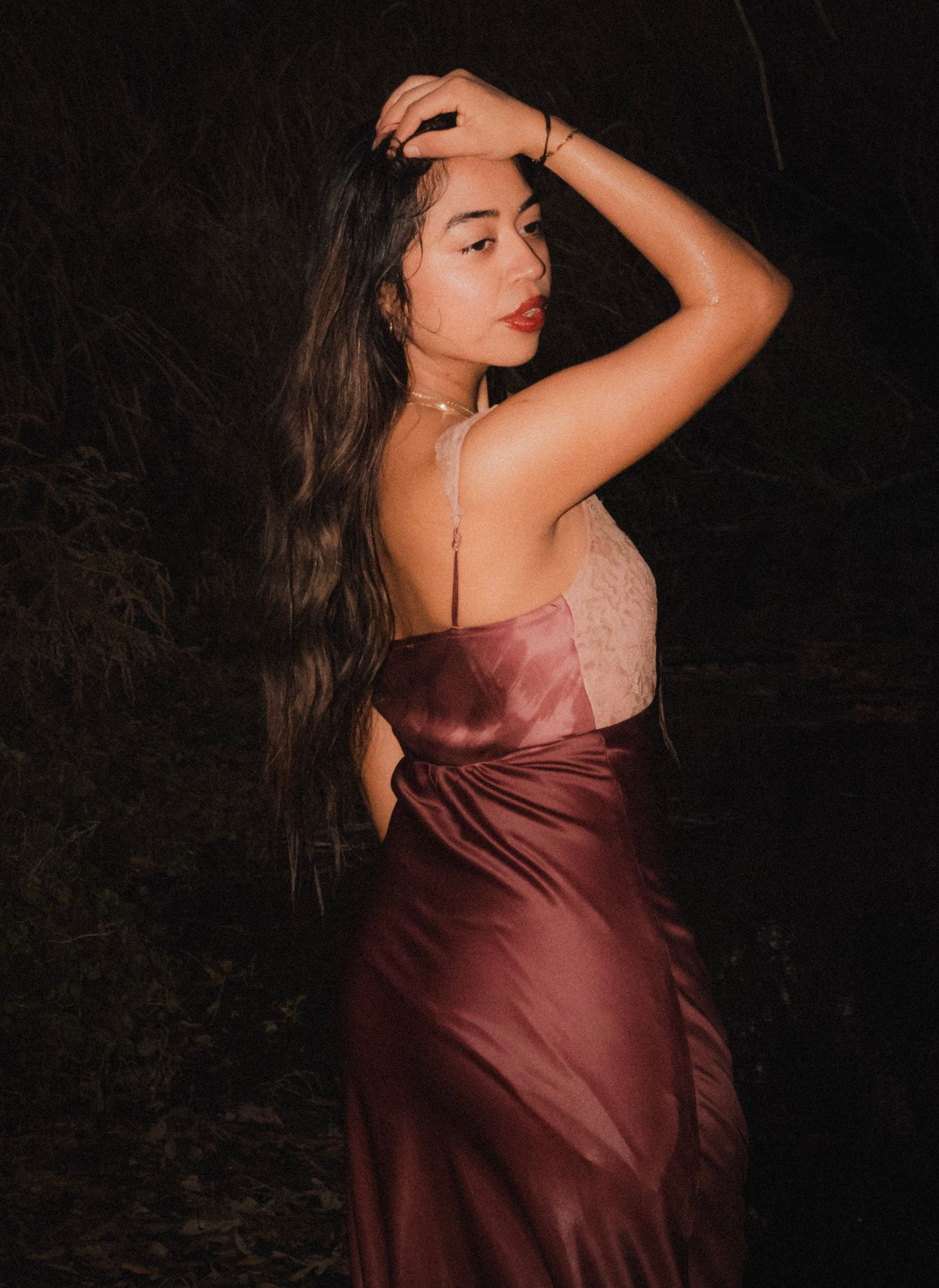 Young woman with long hair in elegant burgundy satin backless gown posing outdoors at night.