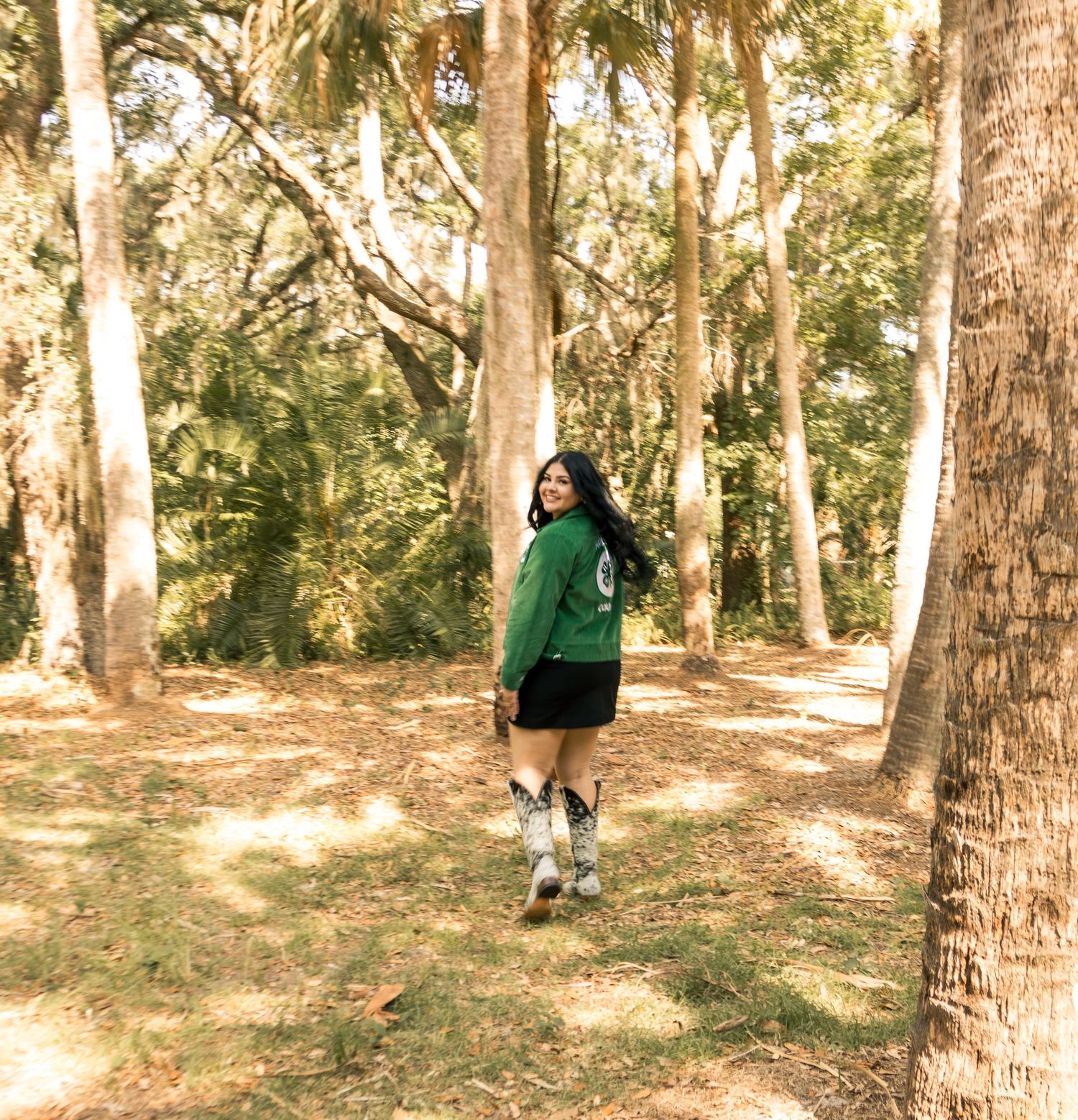 Woman in green jacket and black skirt poses among tall trees in a sunlit forest path.