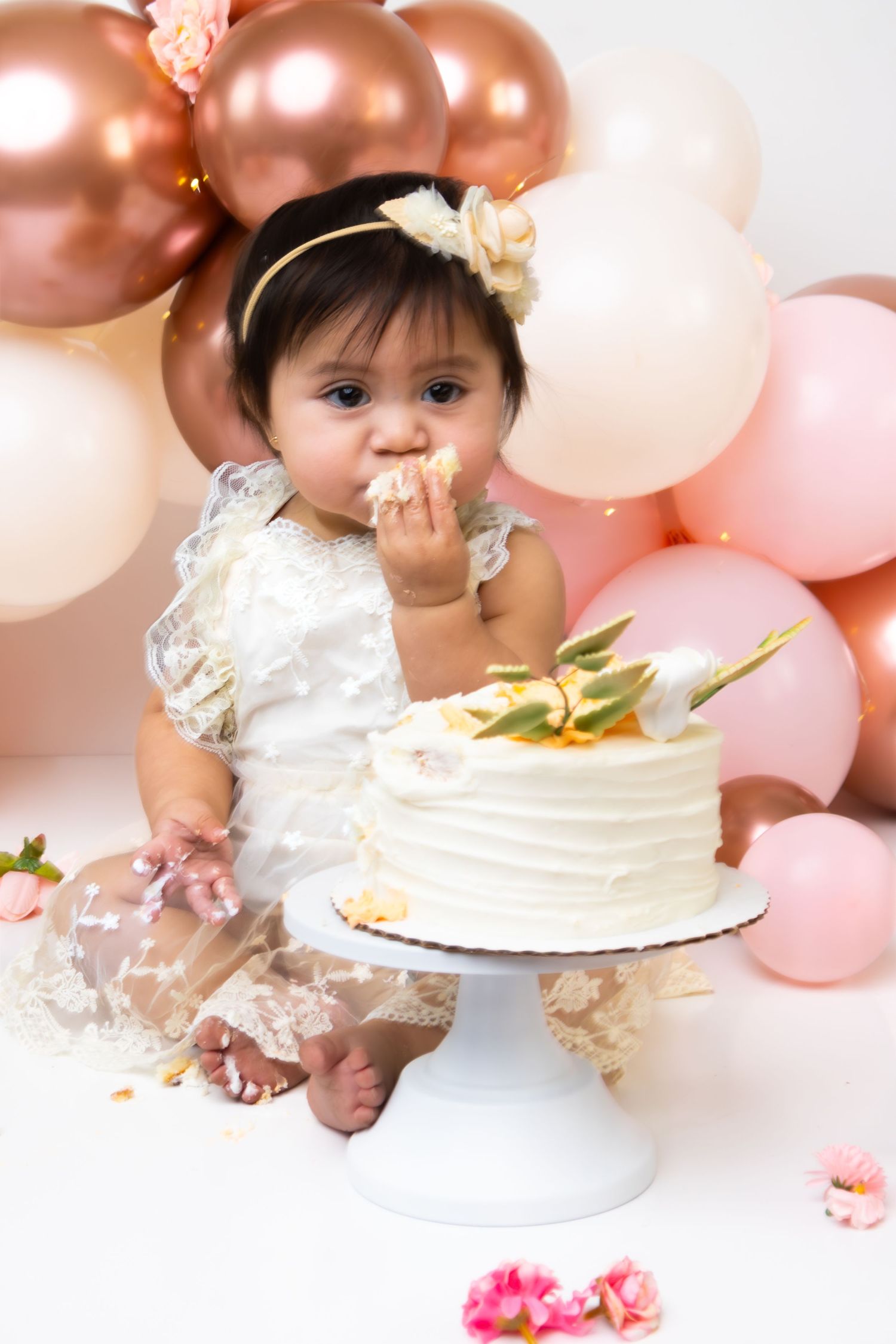Baby girl in white lace dress eating first birthday cake at cake smash with pink and rose gold balloons.