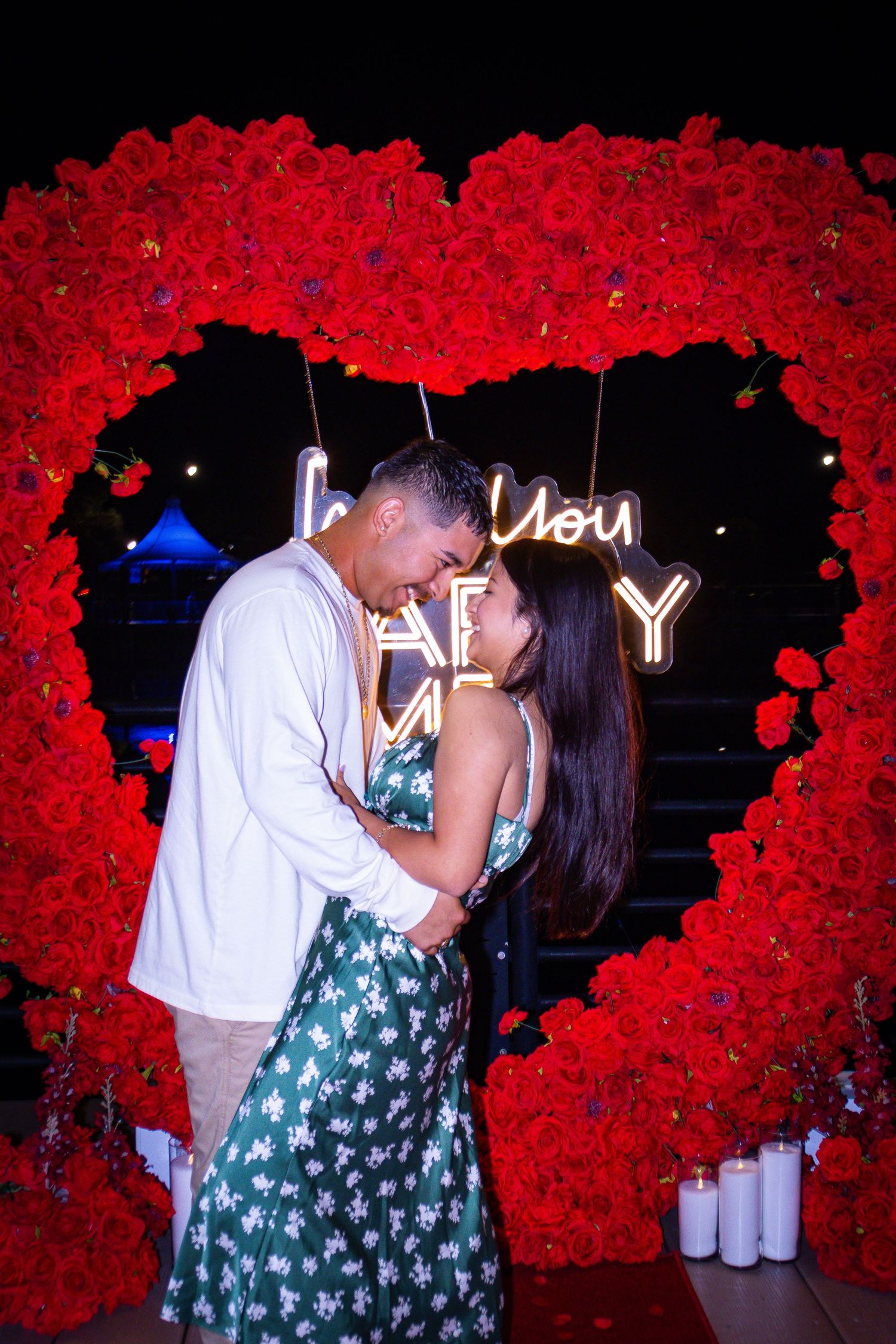 Couple embraces romantically beneath a heart-shaped red rose arch with glowing neon marriage proposal sign.
