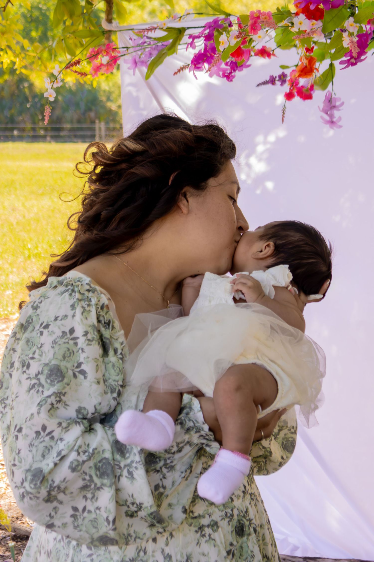 Loving mother kissing baby girl in white tutu dress during outdoor floral themed photo session.