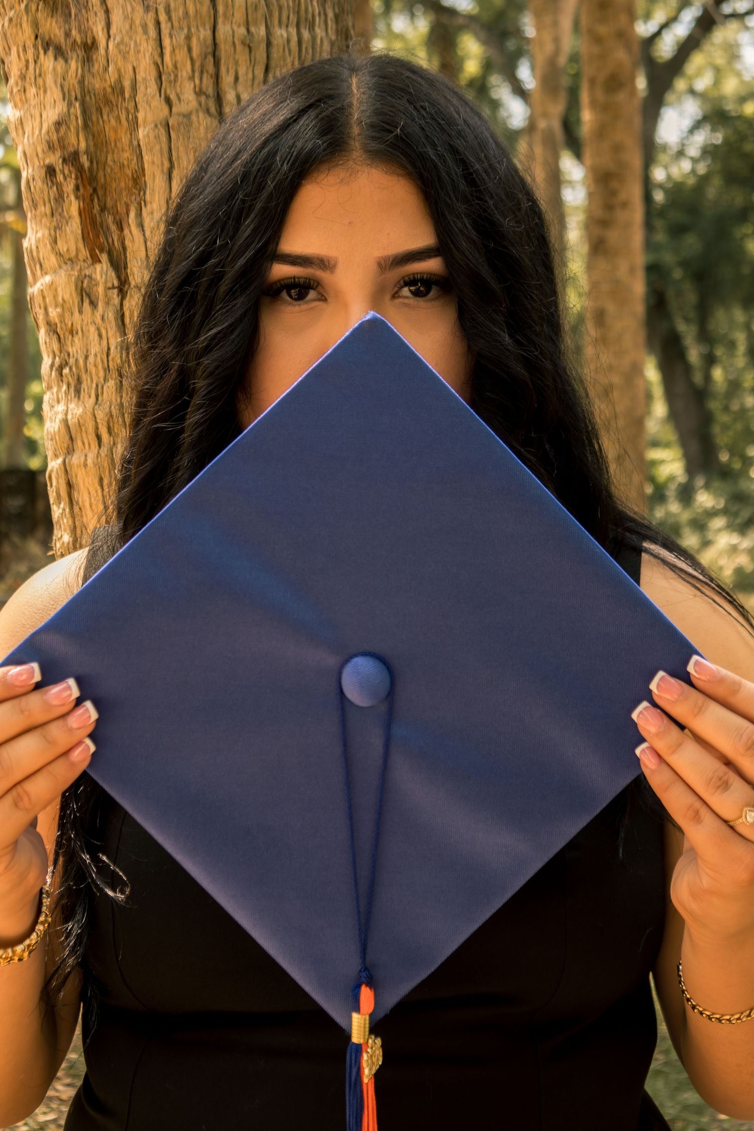 Young woman holding a blue graduation mortarboard cap in front of her face outdoors near trees.