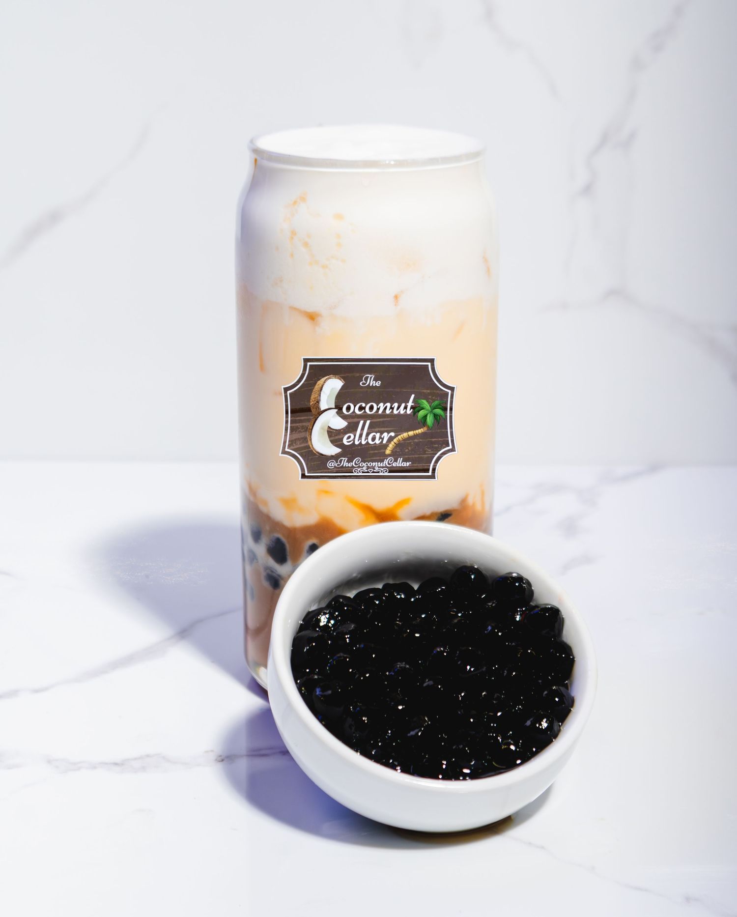 The Coconut Cellar branded boba drink in glass can with bowl of black tapioca pearls on marble surface.