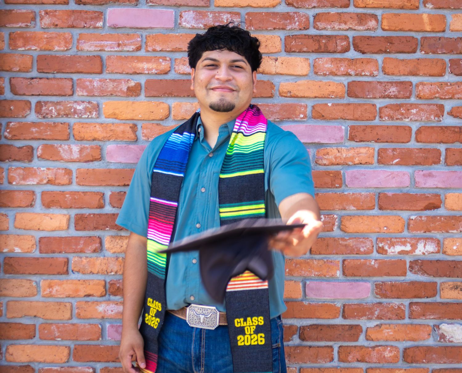 Young man in graduation attire with colorful sash and Class of 2025 stole holding cap before brick wall.