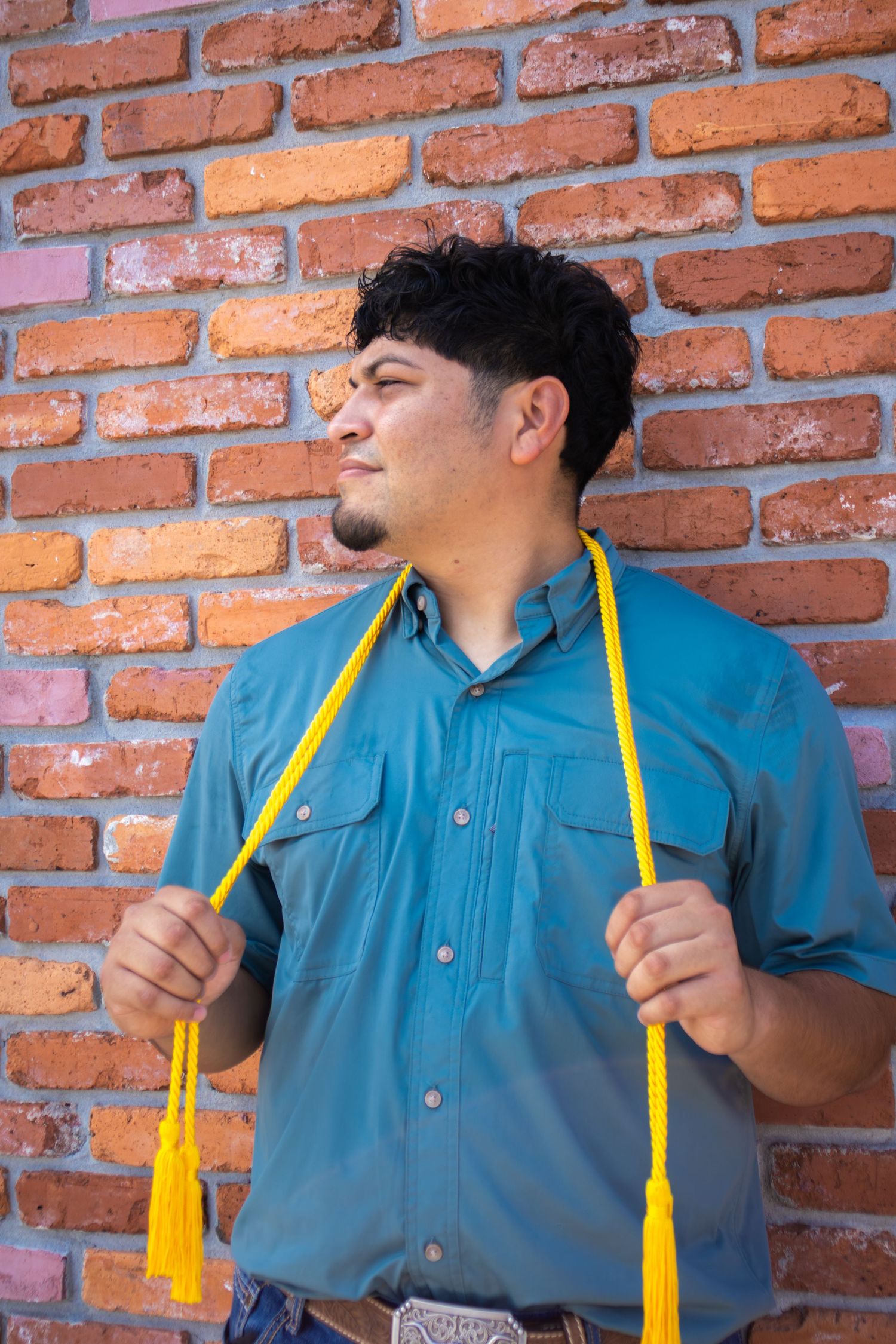 Young Hispanic man in teal shirt holding yellow graduation honor cord against rustic brick wall.