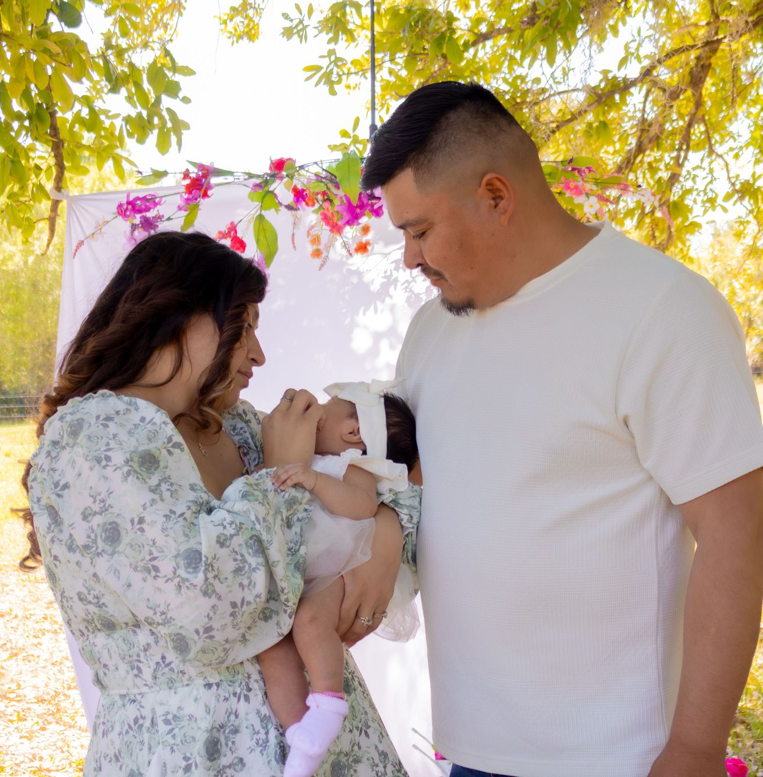 Hispanic couple admires their newborn baby outdoors under flowering bougainvillea tree.