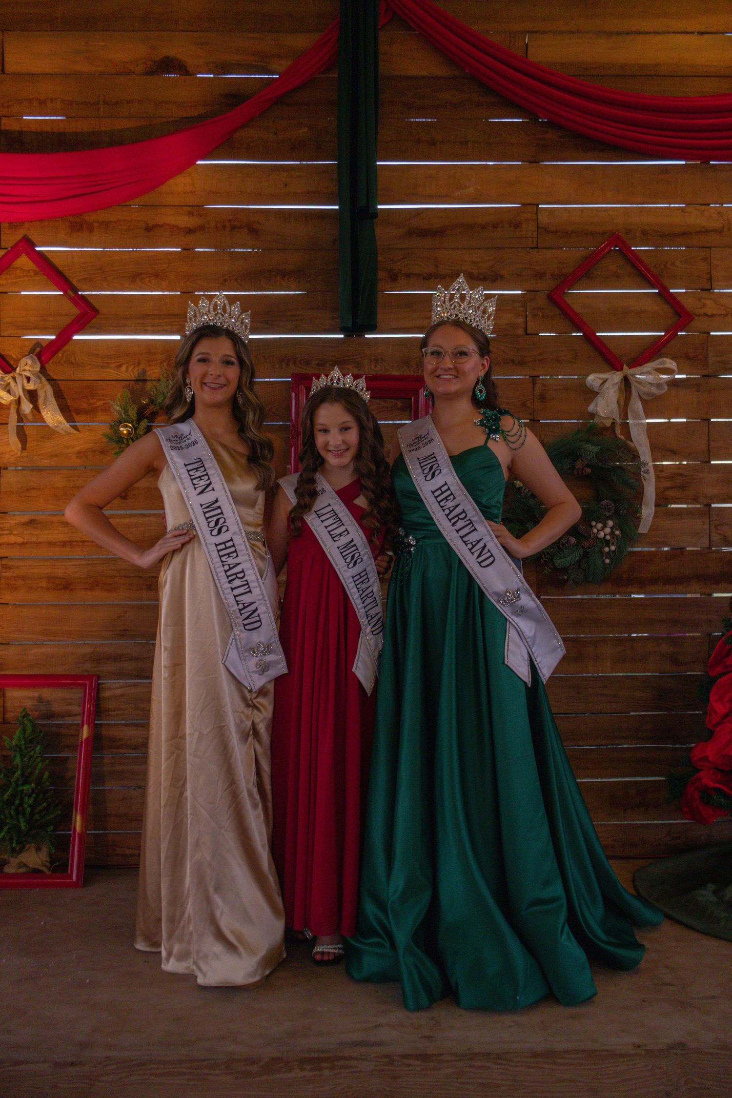 Three beauty queens in elegant gowns and crowns pose wearing Miss Heartland pageant sashes at a rustic Christmas-decorated venue.