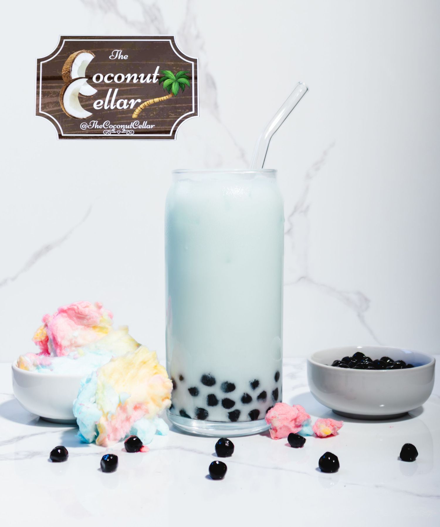 Coconut Cellar bubble tea in a glass cup with boba pearls, cotton candy, and a metal straw on marble background.