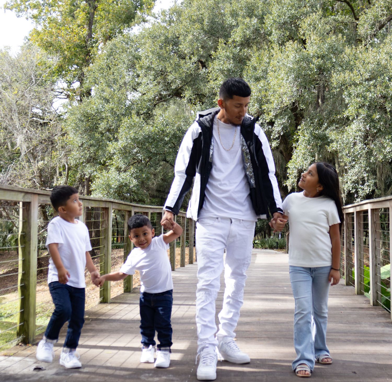 Father walking with three young children on a wooden bridge path surrounded by lush green trees.