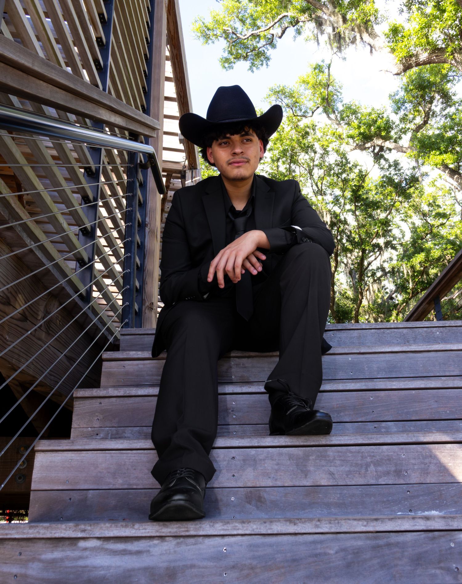 Man in black cowboy hat and suit sitting relaxed on wooden outdoor staircase.