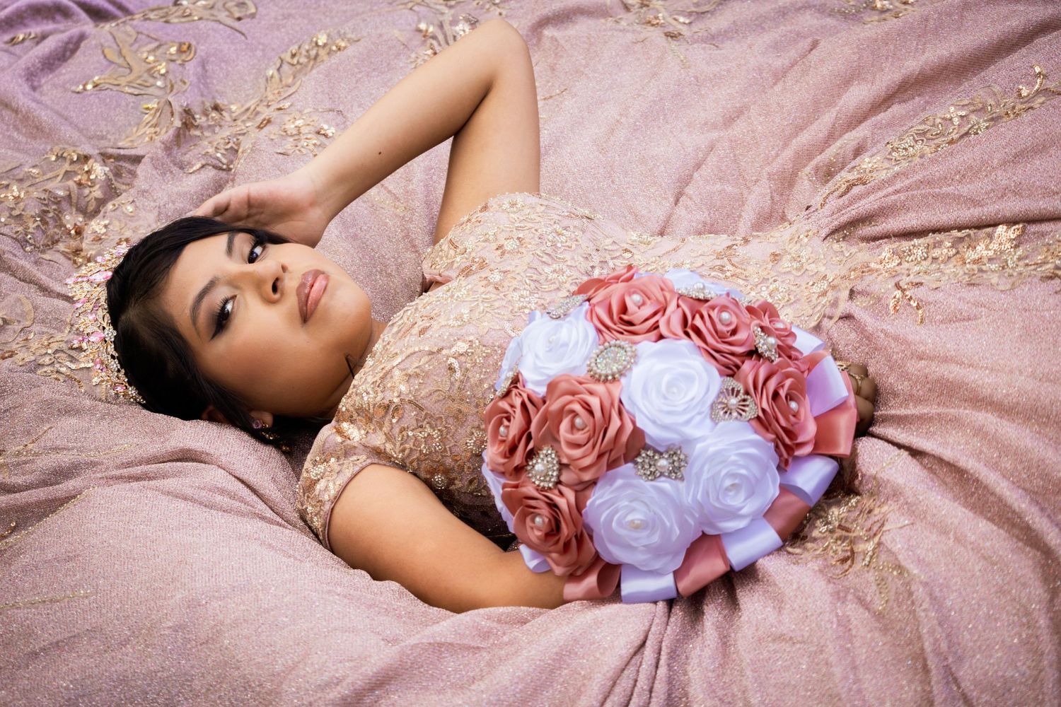 A young woman in a gold quinceañera gown reclines gracefully holding a pink and white rose bouquet.