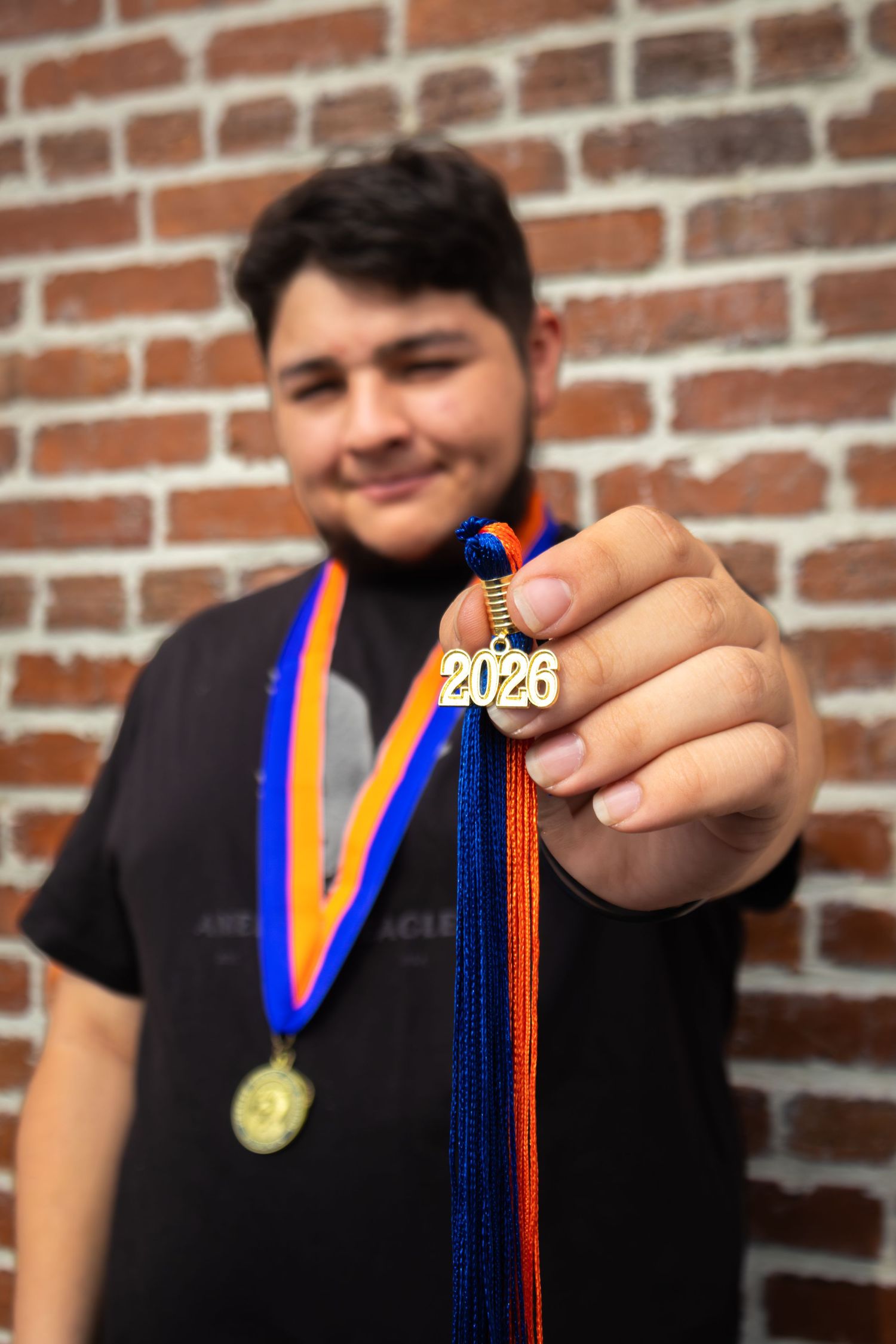 Smiling teen holds a 2026 graduation charm on blue and orange ribbon, wearing a gold medal, against a brick wall.